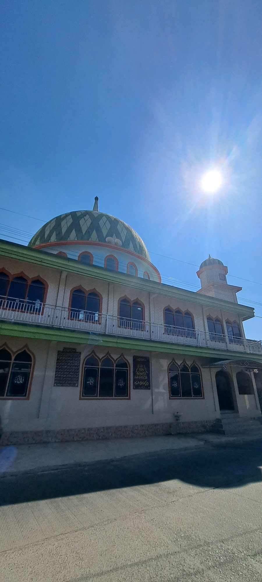The image shows a mosque in the city of Belik, Indonesia, under a clear blue sky with the sun shining brightly and casting a lens flare. The mosque has a prominent green and orange checkered dome and a smaller minaret. The building features multiple stories with balconies and windows. Arabic calligraphy is visible on the walls of the mosque, specifically on plaques and within some of the windows. The foreground shows a paved street with shadows cast by the mosque and possibly other unseen structures. There are no people visible in the image.