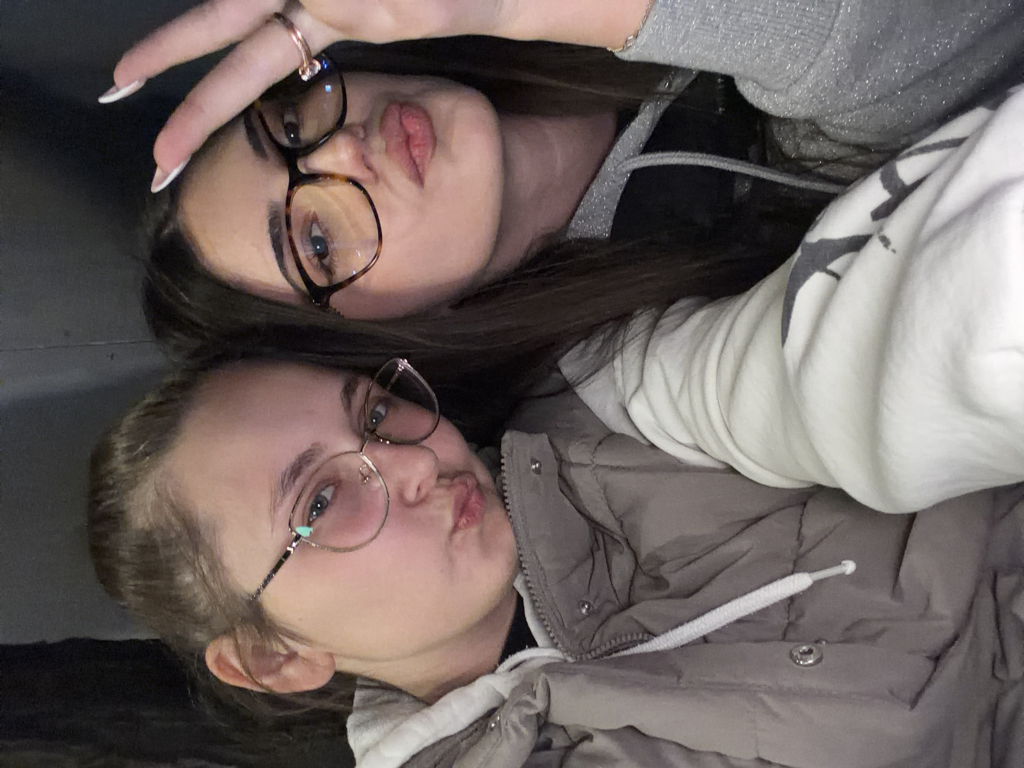 Two young women are posing for a selfie indoors. The woman on top wears brown-framed glasses, a gray hoodie, and has long brown hair. She is making a peace sign with her right hand. She has her lips puckered. The woman below has blonde hair tied back in a ponytail, glasses with metal frames, and wears a gray puffer jacket with a white hoodie underneath. She is also puckering her lips. The location is in Montana, Bulgaria, because the question specified this to be the location.