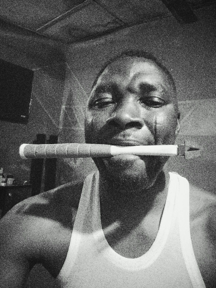A grainy, black and white image displays an adult male in an indoor setting within Gawuna, Nigeria. The subject, positioned centrally and facing forward, wears a light-colored tank top. Prominent vertical scars are visible on both cheeks. He holds a slender, approximately 12-inch long object horizontally in his mouth, clamped between his teeth. The object features a white handle section with dark, transverse bands and tapers to a small, triangular fletching-like end on the right. To the upper left of the subject, a wall-mounted television is partially visible, with indistinct objects resting on a surface below it. The ceiling is visible at the top of the frame. The image exhibits significant visual noise.