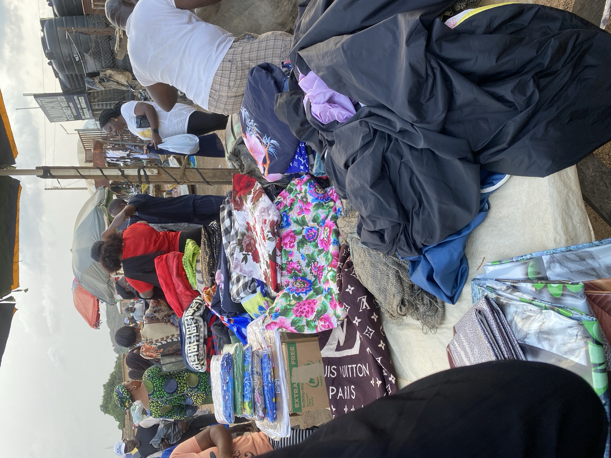 The image depicts an outdoor market scene in Gura Top, Nigeria, with various stalls and a crowd of people. In the foreground, a large pile of black fabric is draped over a stall. To the left, a person wearing a black garment is partially visible. Adjacent to the black fabric, there are stacks of colorful blankets and fabric, including one with a Louis Vuitton pattern. Further to the left, a stack of neatly folded fabric bundles is displayed. In the background, several individuals are seen browsing or tending to stalls, some under umbrellas. The environment appears to be dusty and overcast, typical of an outdoor market. The stalls are made of wood and metal, with tarps and umbrellas providing shade. The overall impression is one of a busy marketplace with a variety of goods for sale.