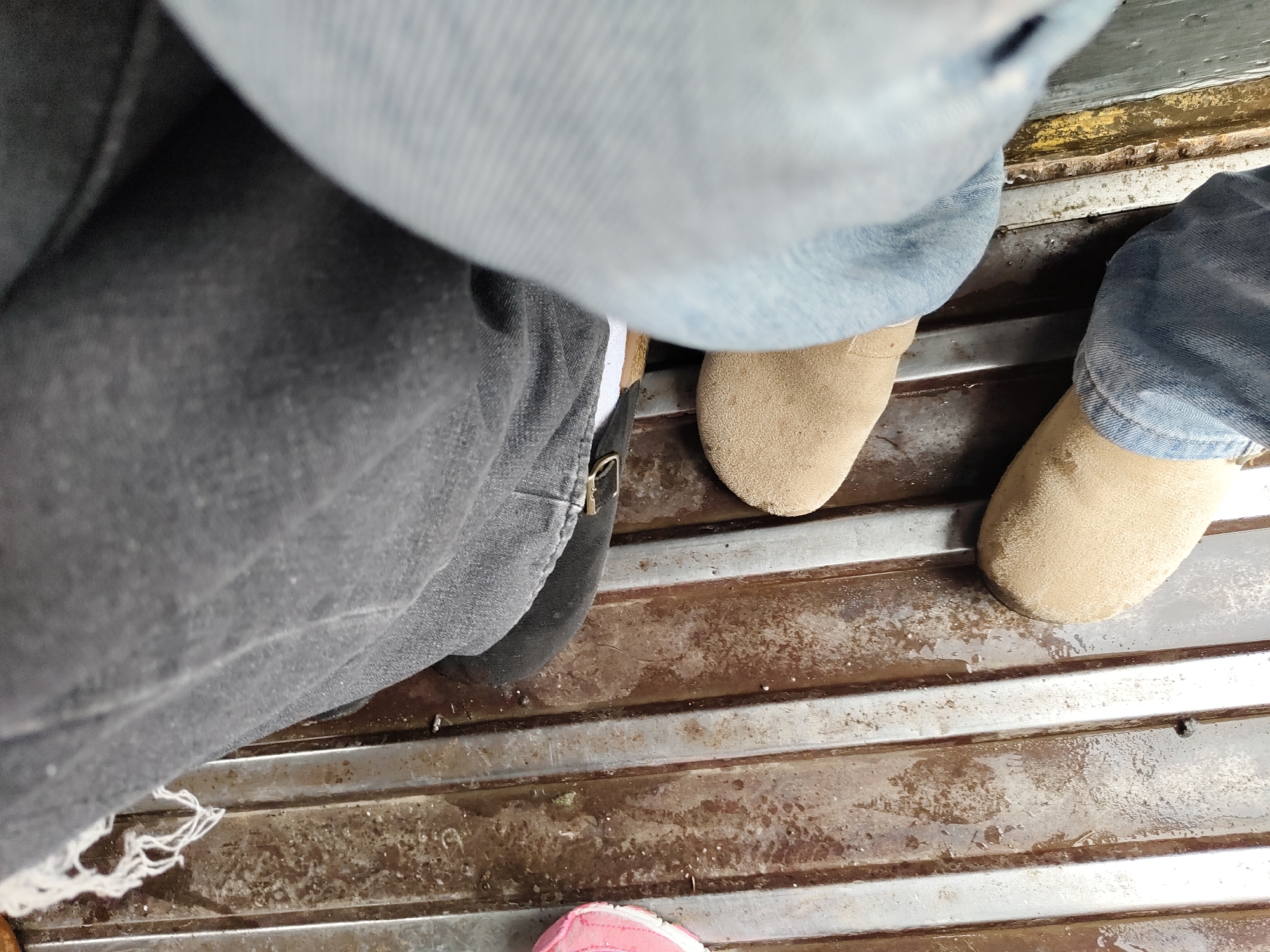 The image shows a close-up, low-angle view of two pairs of legs, one wearing dark grey jeans and the other wearing blue jeans. The legs are positioned on a slatted, weathered surface that appears to be a floor or deck, possibly made of wood and metal. The surface is wet and shows signs of dirt and grime.
On the right side of the frame, one leg is partially visible, wearing a light brown suede shoe. Another light brown suede shoe is visible further to the right. The blue jeans are creased and appear to be made of denim.
On the left side, the dark grey jeans are also creased, with a visible white thread dangling from the hem of one pant leg. A portion of a dark shoe or boot is visible beneath the grey jeans. A pink shoe with white trim is visible at the very bottom of the frame, suggesting a third person is present.
The overall scene suggests a casual setting, possibly an outdoor area or a vehicle interior, with the wet surface indicating recent rain or a damp environment. The image does not provide any specific landmarks or context that would allow for definitive identification of its location within Davao City, Philippines.