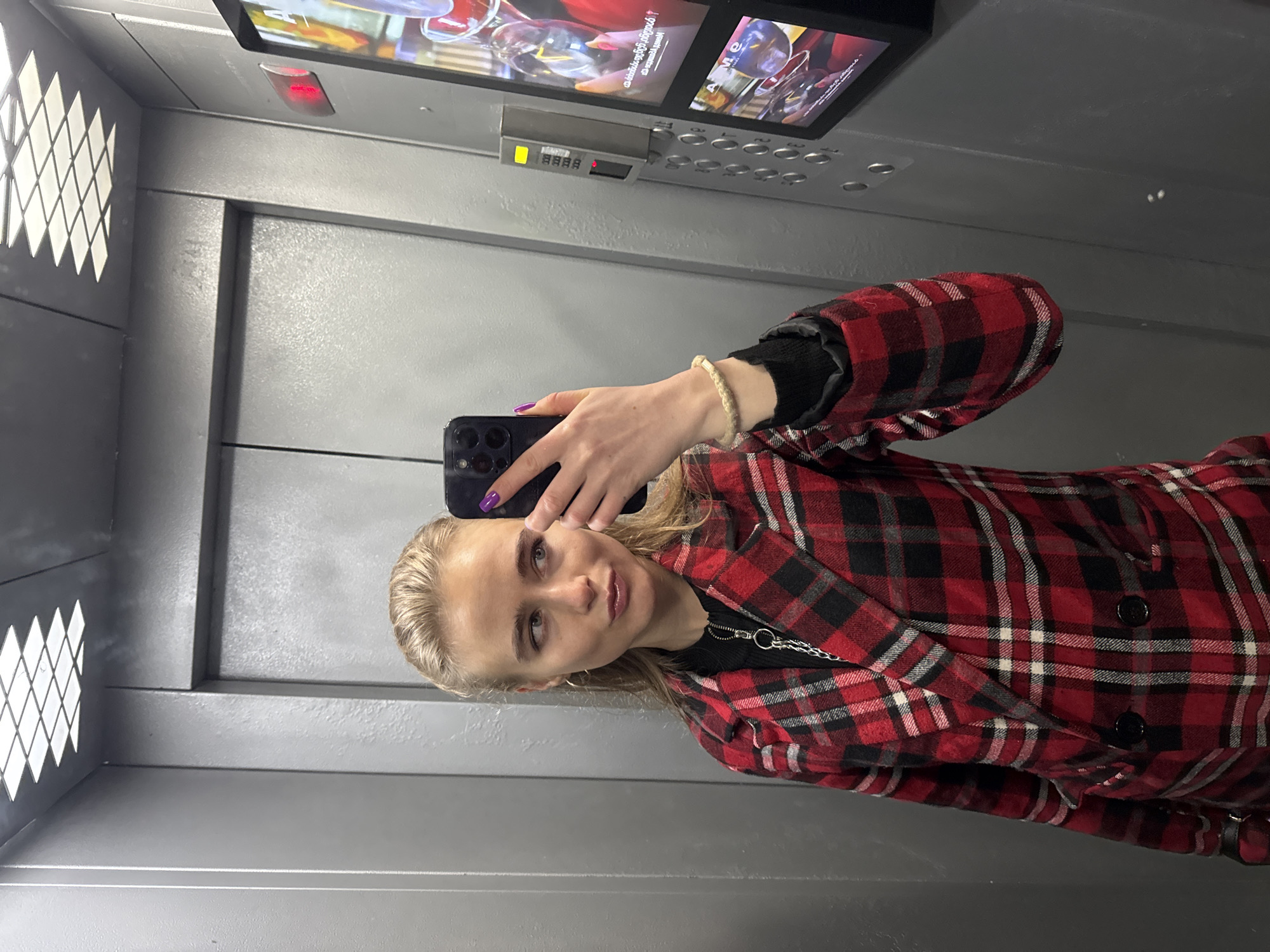A young woman is captured taking a selfie inside an elevator. She has blonde hair and a fair complexion, looking towards the camera with a subtle smile. She is dressed in a red and black plaid jacket worn over a black top, with a silver chain visible around her neck. Her right hand holds a dark-colored smartphone, used to take the picture, and her fingernails are painted purple. A light-colored braided bracelet is on her left wrist. She is also wearing a red and black plaid slipper, matching her jacket.
The elevator's interior features grey metallic walls. To the left, a decorative panel with a diamond-shaped perforated pattern allows light to pass through. Above the woman, an elevator control panel is visible, equipped with numerous buttons, a digital display showing "LL", and a red indicator light. Adjacent to the control panel, a screen displays what appears to be media content or an advertisement, with the text "AMO" partially readable. The lighting within the elevator seems artificial. No specific time of day or outdoor weather conditions can be determined.