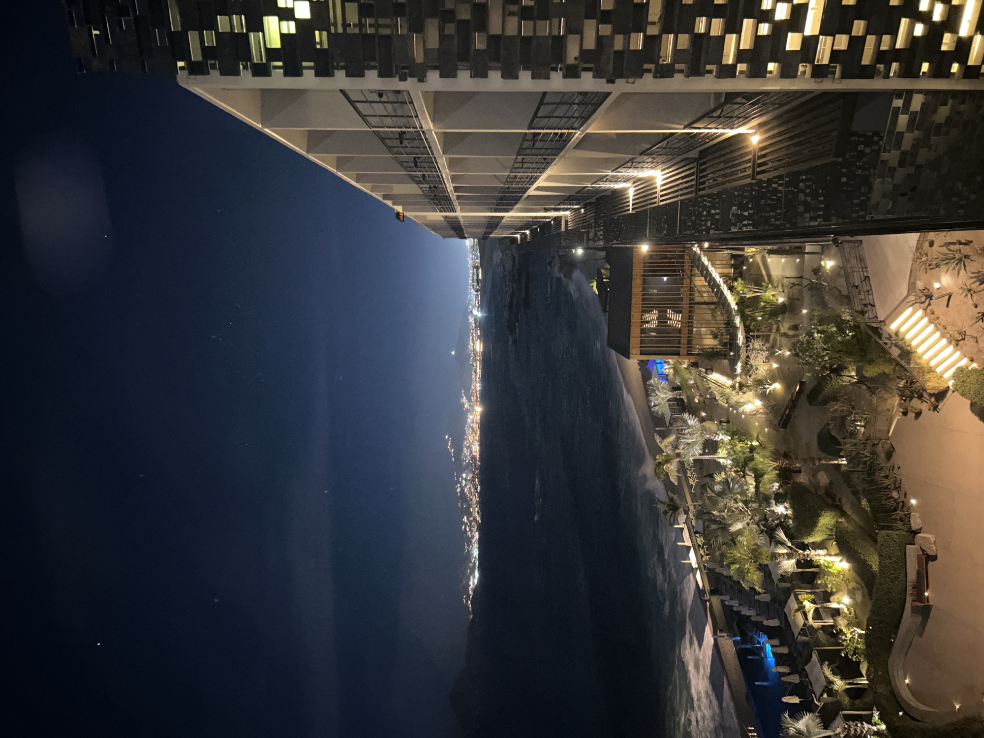 The image depicts a night scene in Cabo San Lucas, Mexico. In the foreground, a multi-story building with illuminated windows is seen from an elevated perspective. The building's modern architecture is visible, with a patterned facade and structural elements. Below, a landscaped area featuring illuminated pathways, palm trees, and other vegetation is present. A body of water, presumably the ocean, separates the illuminated resort area from a coastline dotted with city lights. The night sky is dark, with a few visible stars. The overall scene suggests a high-end resort or hotel overlooking the coastline of Cabo San Lucas.