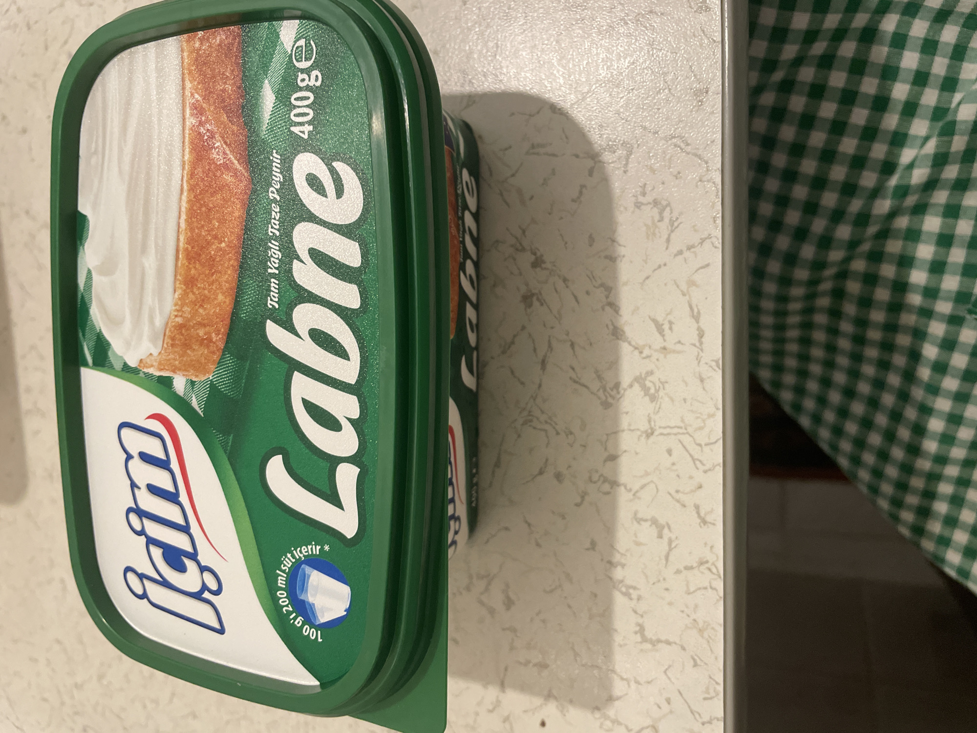 The image shows a close-up view of a green plastic tub of "İçim Labne" cheese, a Turkish brand. The tub is partially opened, revealing a slice of bread topped with white labne cheese. The front label features the brand logo "İçim" in blue and red lettering, and the product name "Labne" in large white letters. Text on the label indicates "Tam Yağlı Taze Peynir" (Full-fat fresh cheese) and "400g". A small graphic of two milk glasses is also visible, with text stating "100 g'ı 200 ml süt içerir" (100g contains 200ml milk). The tub is placed on a speckled countertop, with a green and white checkered fabric visible in the lower part of the frame, suggesting it is on a table or shelf. The background is blurred but appears to be an indoor setting. The image is taken from a slightly elevated angle. There is no information in the image that could indicate its location in Kadıköy, Turkey.