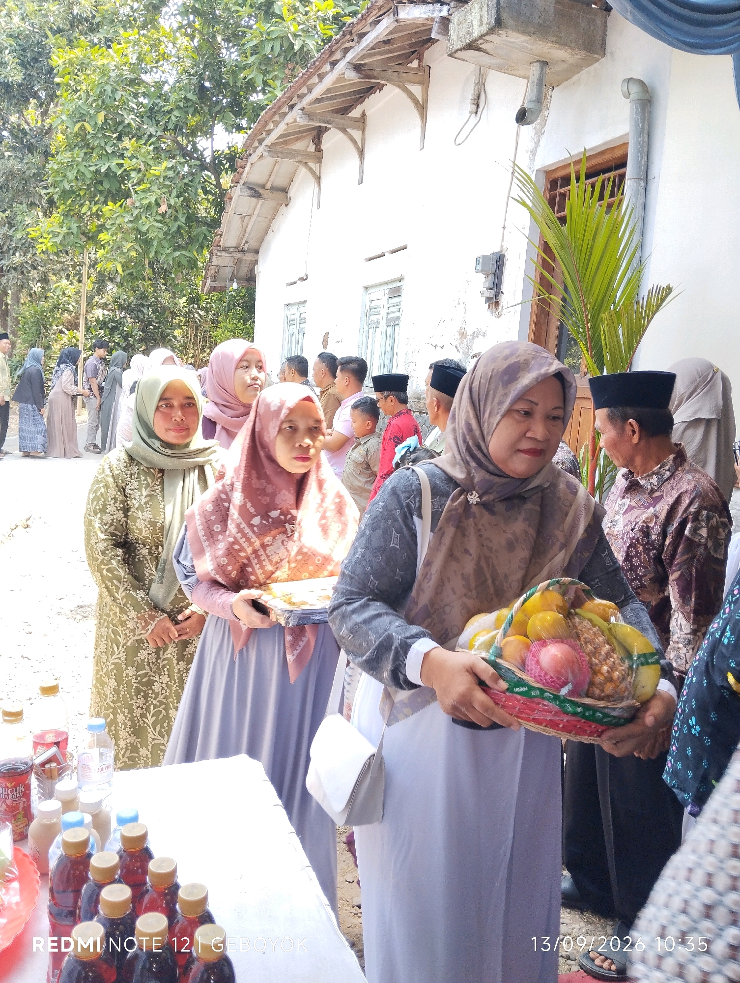 An outdoor scene in Kuwukan, Indonesia, captures a gathering of people, likely for a community event. In the foreground, a woman in a patterned grey hijab and a white collared shirt holds a woven basket overflowing with various fruits, including bananas, oranges, and what appears to be a pineapple. She is dressed in a long, light purple abaya. To her left, another woman in a light green hijab and a green patterned dress stands, also looking forward. Behind them, several other individuals are visible, some in traditional attire. To the far left, a table is laden with numerous bottles of dark liquid, likely beverages, and some small containers. The background features a white building with a textured roof and windows, suggesting a residential or community structure. Trees and foliage are present, indicating a rural or semi-rural setting. The lighting suggests it is daytime.