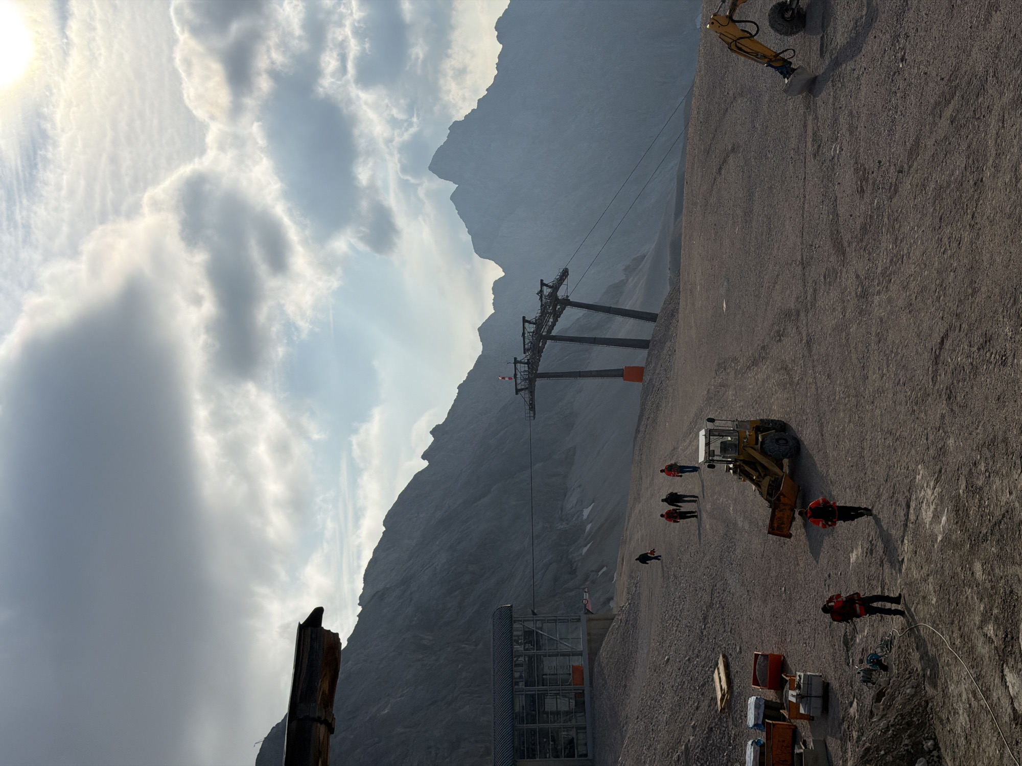 The image depicts a mountainous, construction-site environment, likely at the Zugspitze, Germany's highest peak, near Garmisch-Partenkirchen. The scene is dominated by a vast, barren, gravelly area with several large construction elements, including a cable car station and support towers. The weather appears overcast with patches of sunlight breaking through the clouds, creating dramatic lighting on the rugged mountain backdrop.
In the foreground, a small yellow front-loader bulldozer is parked on the gravel. To the left, several orange construction bins are stacked, and a pallet of what appears to be concrete blocks sits nearby. A person in a red and black safety vest and hard hat stands near some coiled electrical cable, suggesting ongoing work. Further to the left, another person in a similar vest is visible.
In the middle ground, a group of four individuals are walking away from the camera up a slight incline. Three of them are wearing red and black safety vests. Another solitary figure in dark clothing is further up the slope.
In the background, a large cable car station with a glass facade is visible on the left. A substantial cable car pylon structure with cables stretching across the frame is prominent in the mid-to-background. The imposing, jagged peaks of the surrounding mountains form the dominant backdrop, with their slopes appearing rocky and devoid of significant vegetation. The ground throughout the scene is covered in loose gravel and dirt, indicative of a construction or maintenance project in progress at a high altitude. The overall impression is one of a remote, functional, and somewhat harsh, but naturally spectacular, location undergoing development or repair.