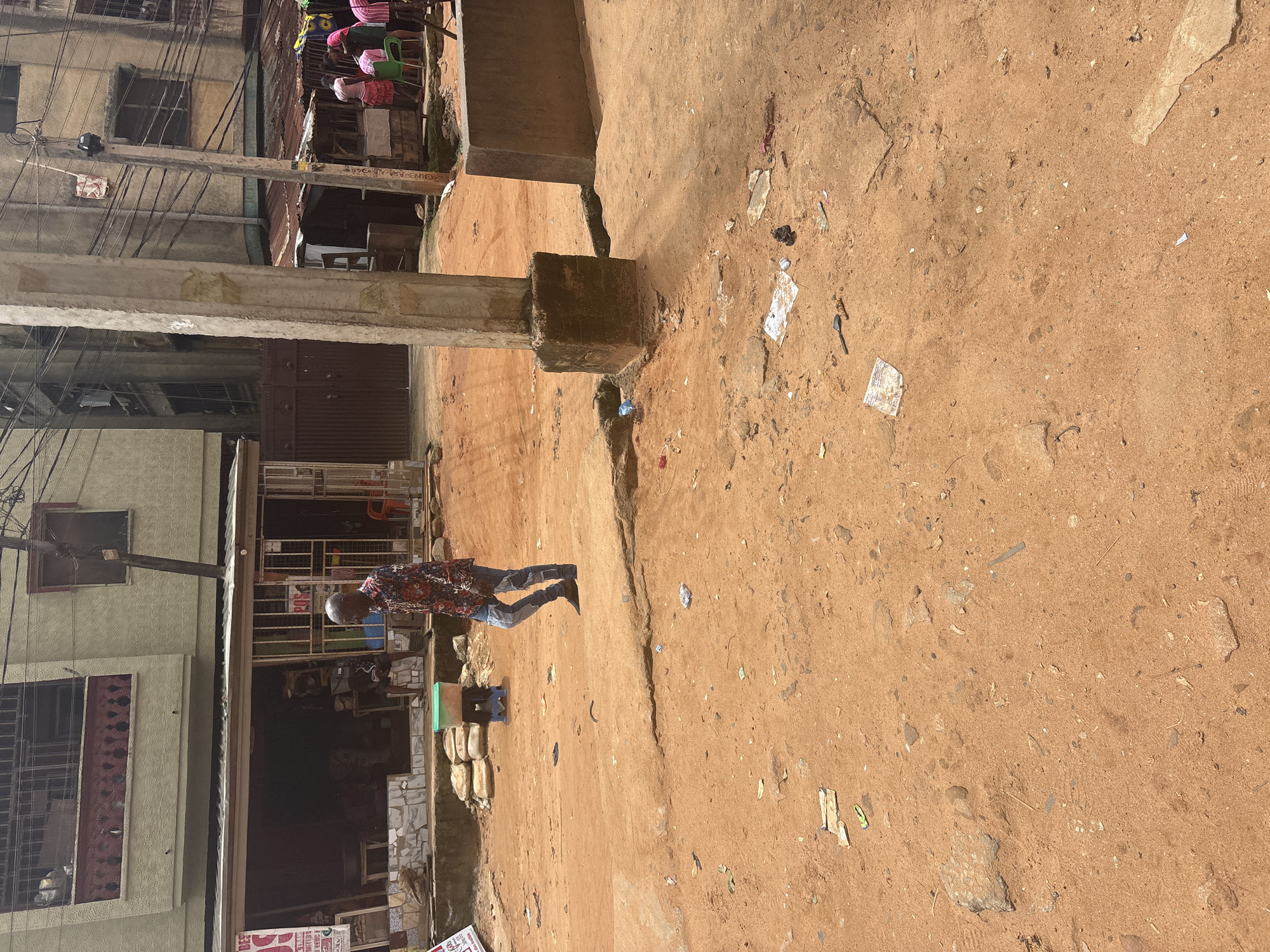 The image shows a man walking on a dirt road in Nkpor, Nigeria. The man is wearing a colorful shirt and jeans. He is walking towards the right side of the image. In the background, there are buildings and shops. The road is dusty and there is some trash on the ground. The environment appears to be an urban setting with commercial activity. There are some people visible in the background near the shops.