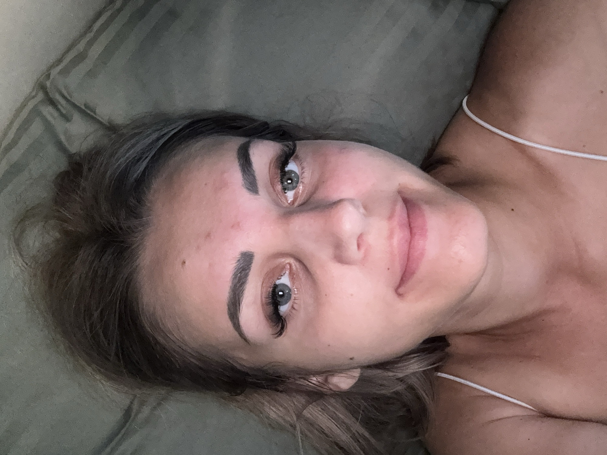 A close-up, overhead shot shows a young woman lying down, looking directly at the camera. Her head is slightly tilted, and her hair is pulled back, with some strands falling loose around her face. She has light-colored eyes, prominent eyelashes, and her eyebrows are dark and well-defined. Her skin appears to have some blemishes, and her lips are slightly parted in a subtle smile. She is wearing a white tank top with thin straps. The background consists of a textured, dark green surface, possibly a pillow or blanket. The scene is dimly lit, creating soft shadows and highlighting the contours of her face. No specific location in Yuzhno-Sakhalinsk, Russia can be determined from the image.