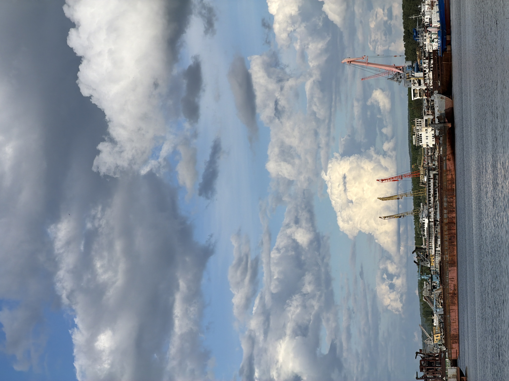 The image depicts a view of the water with docked barges and cranes against a backdrop of a cloudy sky. The sky is filled with varying shades of grey and white clouds. Several large barges are docked along the water’s edge, with cranes interspersed amongst them. Trees can be seen in the background along the shoreline. The scene is located in Chaikovsky, Russia.