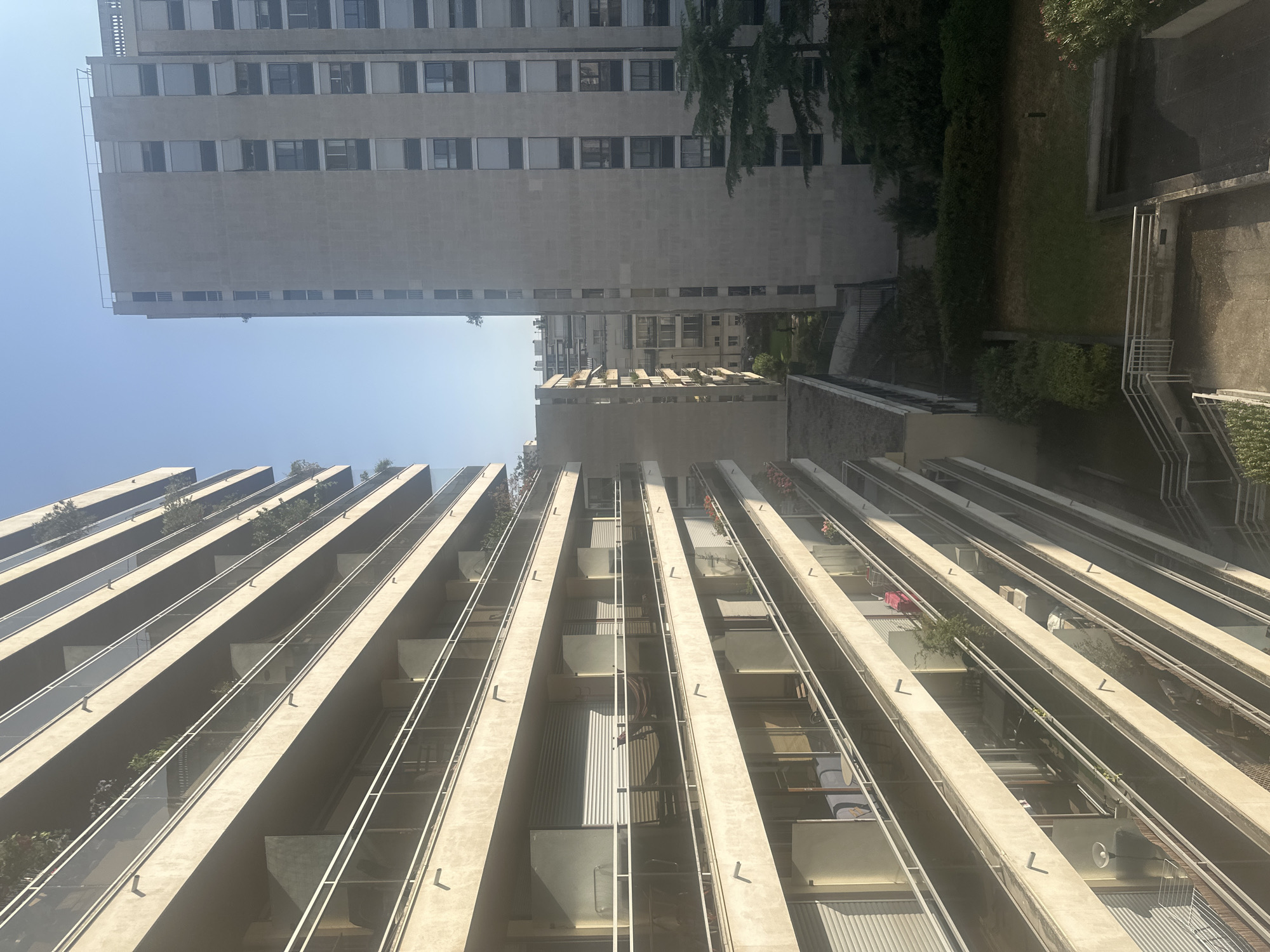 The image shows an outdoor view of residential buildings in Paris, France. In the foreground, a modern apartment building with numerous balconies is visible. The balconies have glass railings and are furnished with various items, including plants, chairs, tables, and laundry. Further in the background, another tall, grey residential building with many windows and closed shutters stands. Between these buildings, there is a courtyard with green spaces and trees. The sky is clear and blue, suggesting a sunny day. The perspective is from a higher vantage point, looking down and across at the buildings and the courtyard.