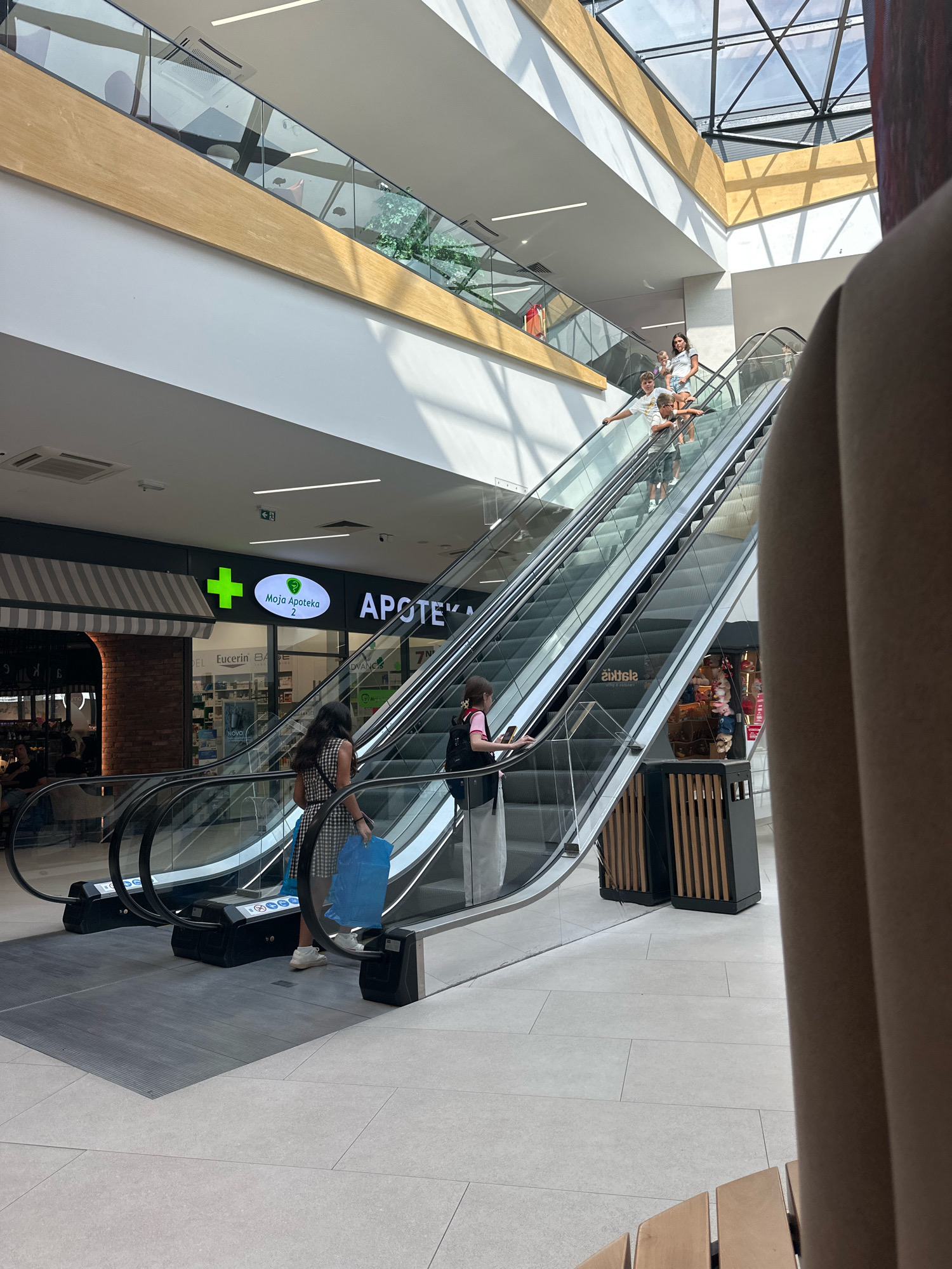 The image depicts the interior of a modern shopping mall with natural light filtering in through a large skylight. Several people are visible on escalators. On the lower escalator, a woman in a checkered dress and white sneakers, carrying a bright blue shopping bag, is ascending. To her right, another person, possibly a woman wearing a pink top and dark pants, is also ascending the escalator. Further up on the same escalator, a group of children are seen ascending, with two boys in the foreground and two girls behind them. On the upper escalator, another group of people are ascending, with the figures less distinct.
The environment features polished light-colored tiled flooring, glass railings along the escalators, and wooden architectural elements, including a slatted partition to the right. Above the escalators, signs for businesses are visible, including "MOJA APOTEKA 2" and "APOTEKA". The mall's infrastructure, such as ceiling panels and air conditioning units, is also apparent. The overall impression is a busy, contemporary commercial space. This location is identified as the shopping mall in the city of Budva, Montenegro.