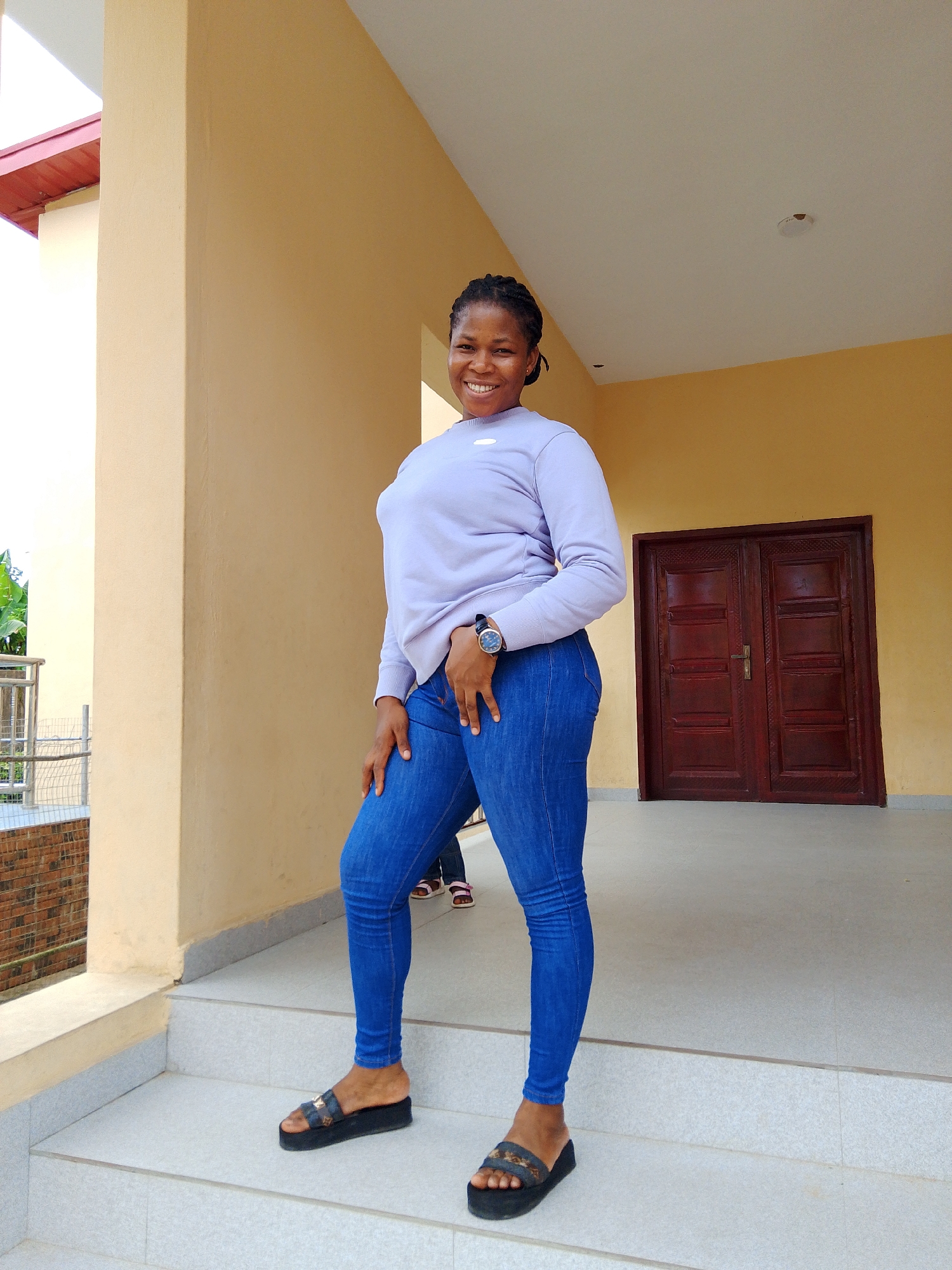 In Yenegoa, Nigeria, a young woman stands on a set of outdoor stairs, posing for a photo. She is wearing a light purple long-sleeved shirt and form-fitting blue jeans, accessorized with a wristwatch on her left wrist and dark platform sandals. Her hair is braided and pulled back. The background features a building with a cream-colored facade and a large, dark wooden double door. A beige column is to her left, and to her far left, a railing and some greenery are visible. The stairs are made of a light-colored, textured material.