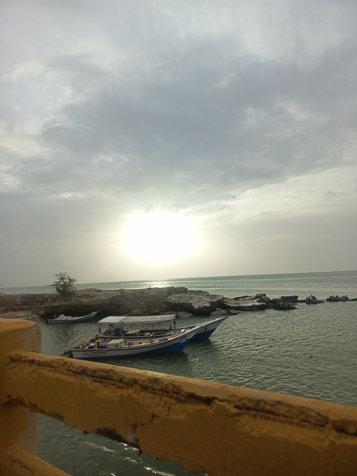 The image shows a coastal scene in Juan Griego, Venezuela. The sky is overcast with a bright, obscured sun. The foreground features a yellow railing. In the middle ground, several boats are docked in the water near a rocky shoreline. Some birds are perched on the rocks farther out in the water. A small tree grows on the shoreline. The water surface is rippled and reflects the sunlight.