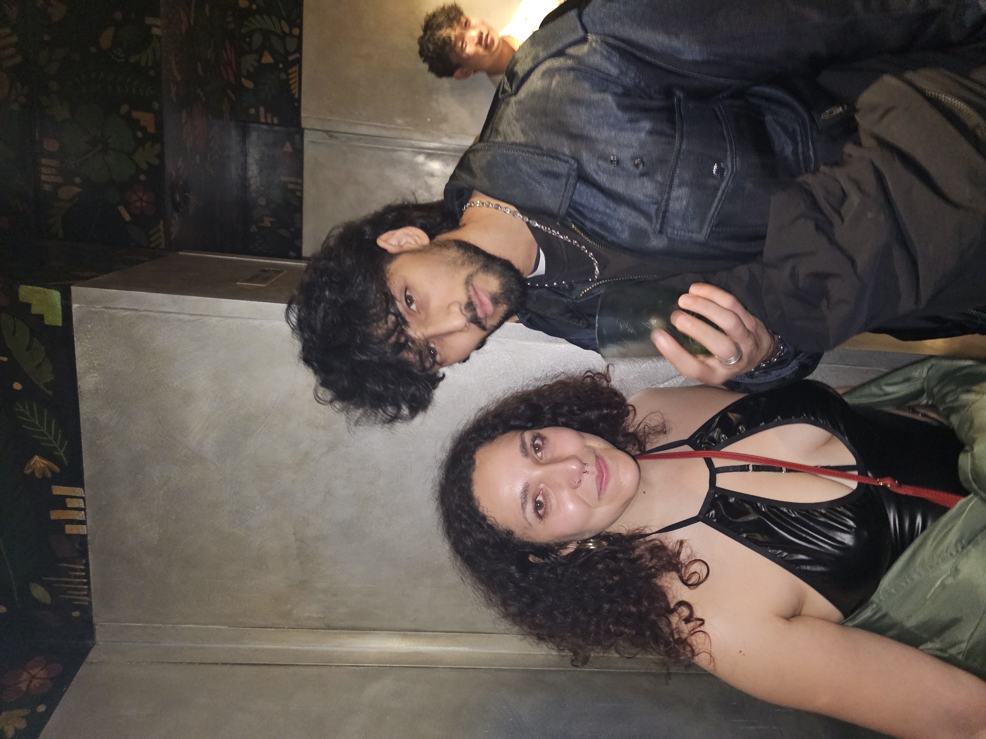 Three people are in an interior space. A woman with curly hair and a black top is positioned in the foreground. Behind her and slightly to the left is a man with curly hair, a beard, and a denim jacket. Further in the background and above the man is another man. The background is dimly lit with a mix of textured walls and patterned wallpaper. The location could not be identified from the visual data as being located in Paris, France.