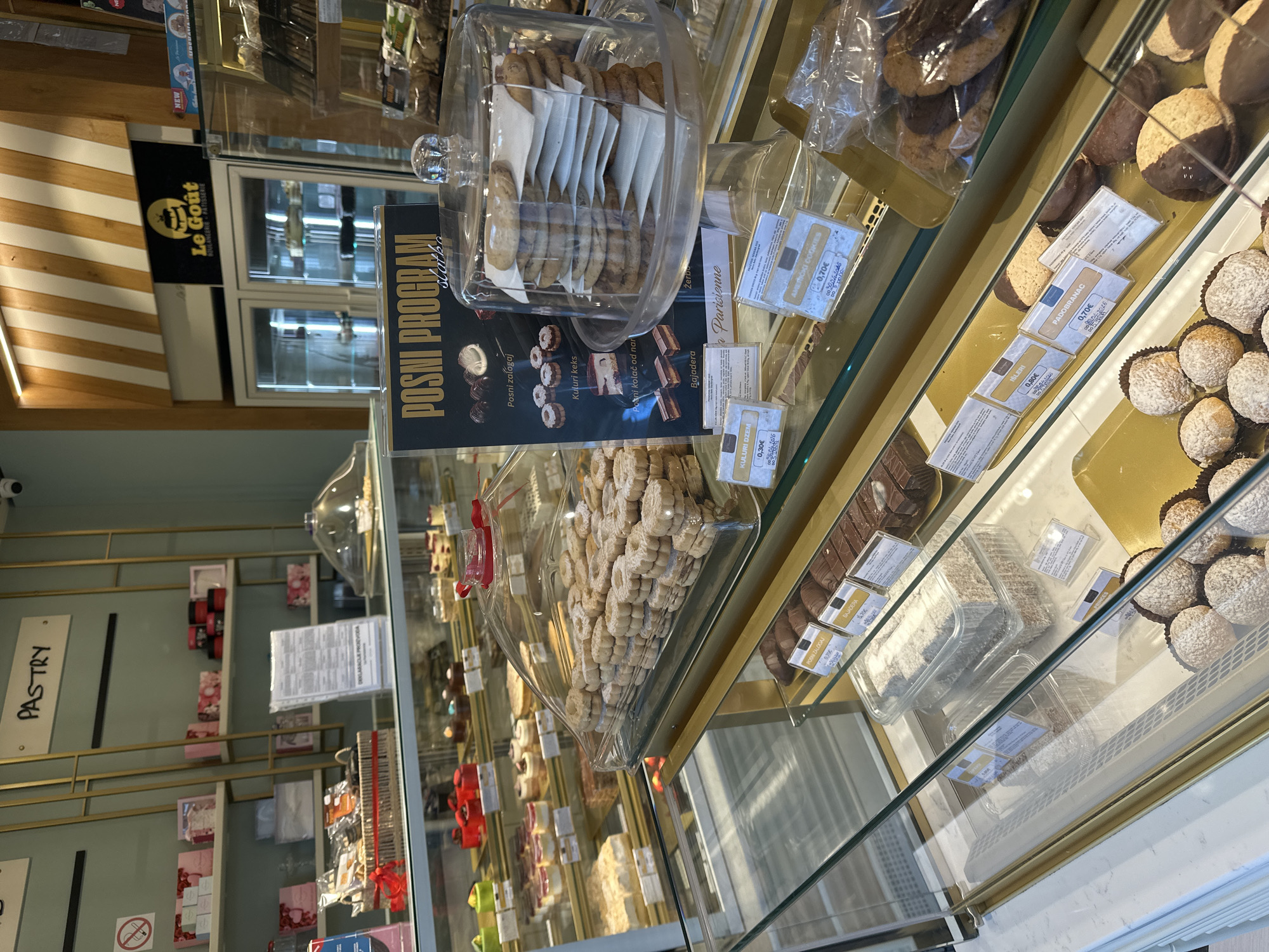 The image depicts a pastry display case inside a bakery, possibly "Le Goût" in Kolašin, Montenegro, based on the sign visible in the background. The case contains various pastries and cookies, including those labeled as part of a "Posni Program" (Lenten program). The pastries are displayed under clear domes and in plastic containers, with price tags visible for items such as "Kuluri dzem" (€0.30), "Žerbo," "Bajadera" (€1.70), "Išleri" (€0.80), "Američki Cookies" (€0.70), and "Padobranac" (€0.70). Other baked goods are visible, like chocolate-covered cookies and coconut-covered treats. Shelves with products are visible in the background of the bakery. A sign reading "PASTRY" hangs to the left.