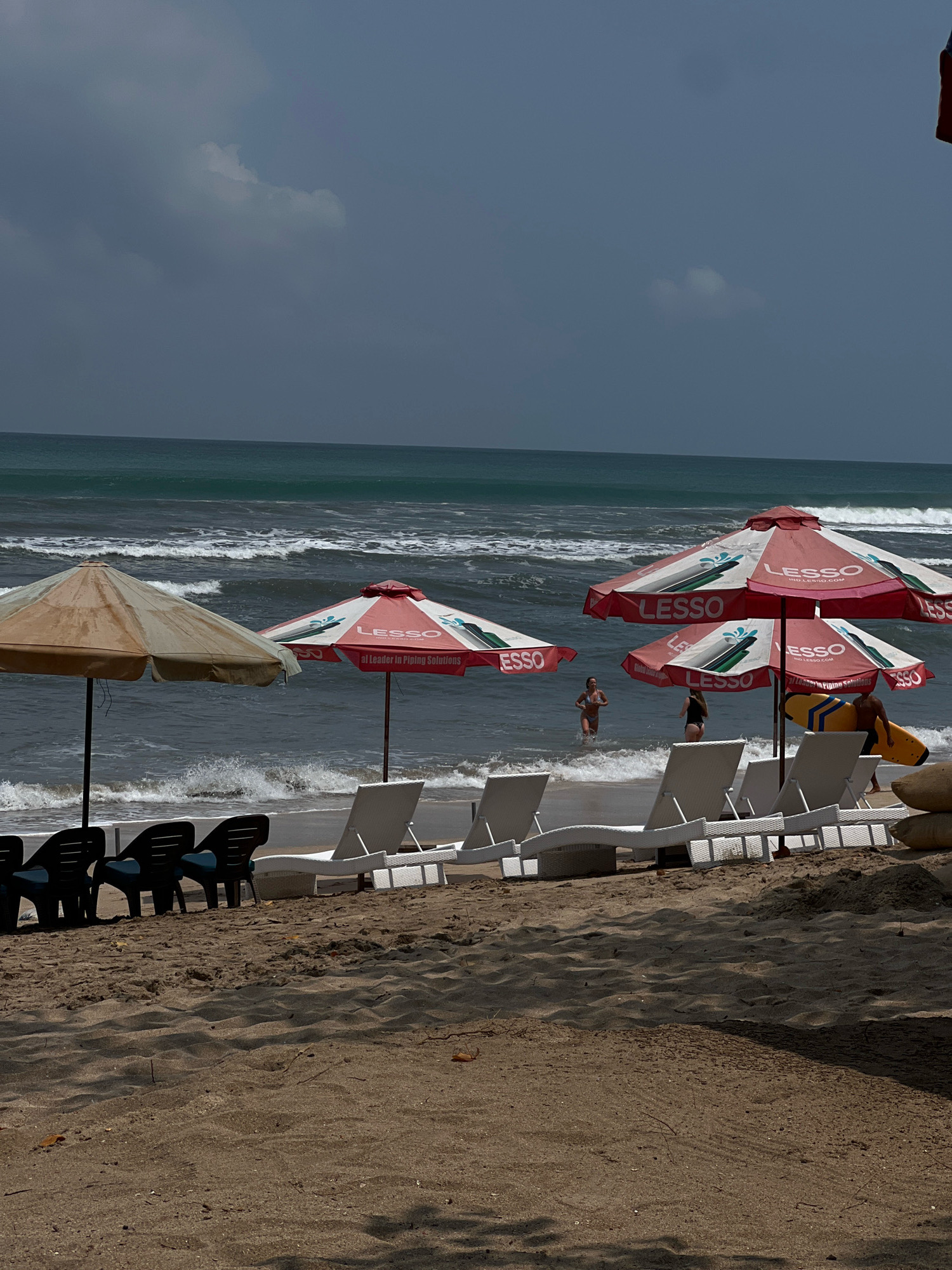A group of beach umbrellas and lounge chairs are set up on a sandy beach in Kuta, Indonesia, with the ocean in the background. The sky is overcast and dark, with some patches of lighter cloud. The ocean has moderate waves with whitecaps.
In the foreground, there are several black plastic chairs lined up, some with blue cushions. Behind them, a row of white lounge chairs are set up on the sand. Two large, round beach umbrellas are prominently displayed. One is a beige, weathered umbrella. The other is a red and white umbrella with the logo "LESSO" and text that reads "al Leader in Piping Solutions". Further back, another red and white umbrella with the "LESSO" logo is visible.
In the ocean, several people are visible. Two women are in the water, one in a blue bikini and the other in a black swimsuit. A man is walking out of the water carrying a yellow and blue surfboard.
The sand on the beach is light brown and appears to be a mix of fine and coarser grains. There are some dried leaves and debris on the sand. In the far right of the image, there are some sandbags. The overall impression is of a public beach with amenities available for visitors, under a somewhat gloomy sky.