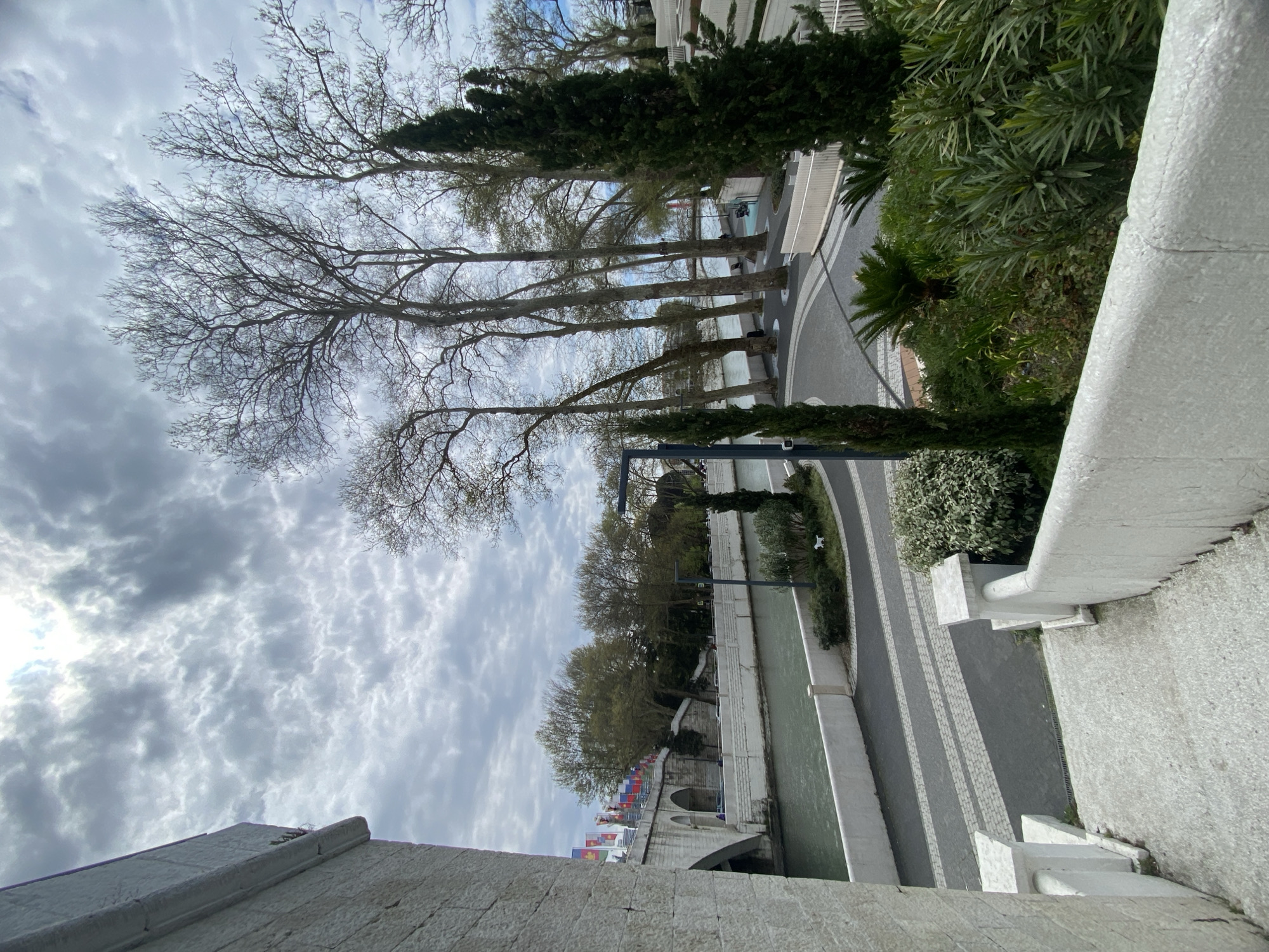 This image captures a serene waterside scene, likely in Sochi, Russia, viewed from an elevated position. The perspective looks down onto a body of water—a river or canal—and across to the opposite bank.
In the foreground, a textured, light-colored stone balustrade or wall runs diagonally, framing the right side of the view. To the right of the water, a paved walkway, characterized by a pattern of dark and light gray interlocking pavers, curves alongside the bank. This path is bordered by lush green landscaping, including various shrubs and palm-like plants. A modern street lamp stands amidst the greenery.
The river itself appears a murky green. On the opposite bank, a prominent stone structure with multiple archways, possibly a historic bridge or a reinforced quay wall, is a key feature. Along the top of this structure, numerous small, colorful flags, which appear to be heraldic or regional banners rather than national flags, are displayed.
Behind the arched structure, a line of tall, bare deciduous trees with intricate branch patterns stands against the sky, suggesting late winter or early spring. Further back, more distant trees and hints of buildings can be seen.
The sky is heavily overcast with a wide expanse of mottled grey and white clouds, indicating a cloudy or diffused light day. There is no direct sunlight. The scene appears devoid of people, and no legible text is visible anywhere in the image. The overall atmosphere is tranquil and calm, showcasing a blend of urban architecture and natural elements.
