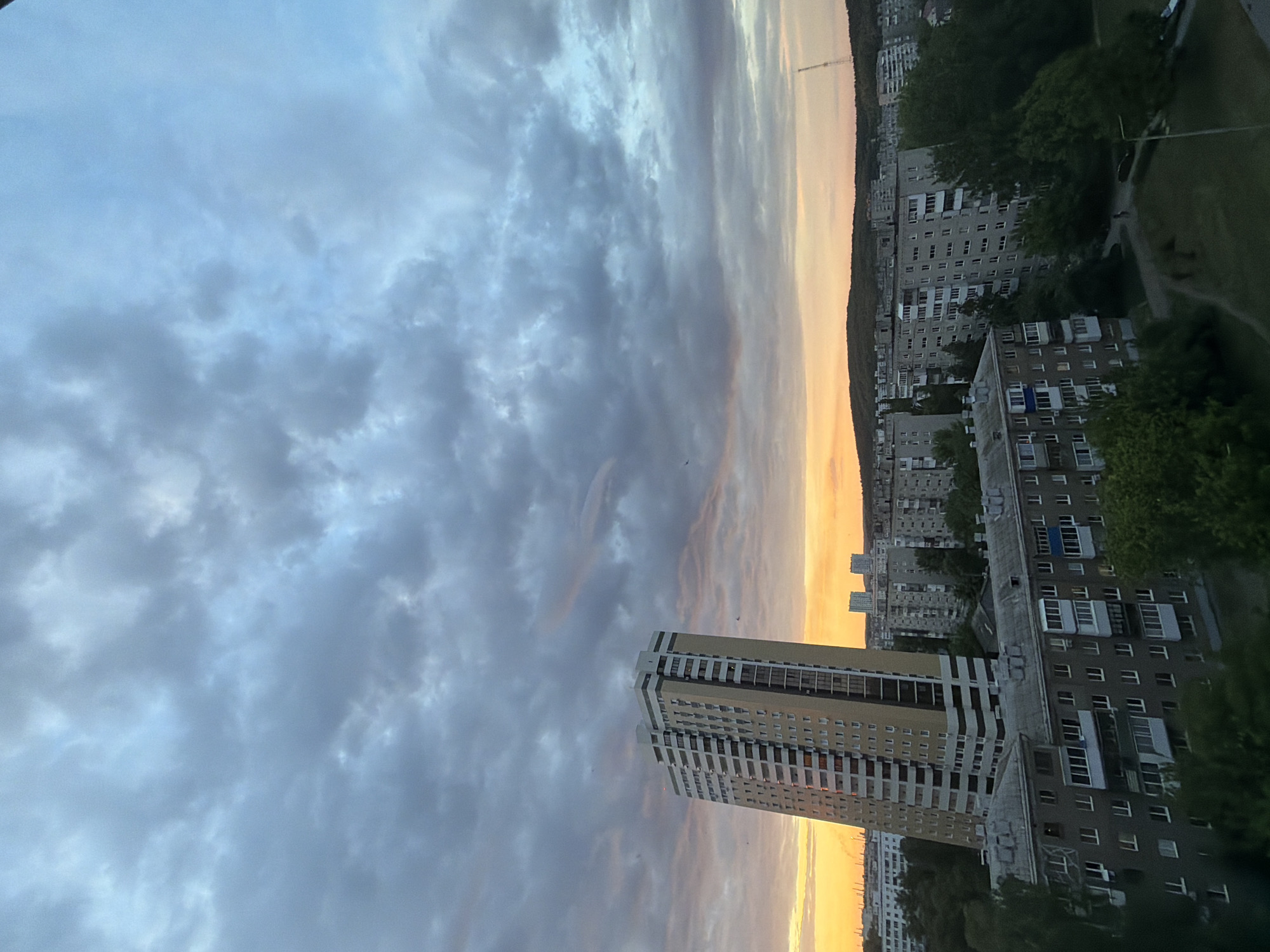 The image, correctly oriented in landscape, presents an urban panorama at dusk or dawn, characterized by a dramatic sky and a dense residential landscape. The expansive sky occupies the upper two-thirds of the frame, filled with a mix of heavy grey and lighter blue-white clouds. Along the horizon, a striking band of golden-orange and soft yellow light indicates the setting or rising sun, contrasting with the cooler tones of the clouds above.
In the mid-ground, a prominent, modern high-rise building with a beige and white exterior stands on the left. Its numerous windows reflect the subdued light of the sky. To its right and extending into the background, a vast array of older, multi-story apartment buildings, predominantly grey and featuring many balconies, fills the cityscape. These structures are nestled among pockets of green trees and foliage, suggesting parks or street-side greenery. In the distant background, a gentle hill or forested area is silhouetted against the horizon, with a slender radio tower faintly visible on the far right.
The scene is tranquil, with no visible people or discernible activity. The soft, directional lighting and color palette strongly suggest either sunrise or sunset, with the rich orange hues leaning towards sunset. The weather appears overcast but calm. There is no visible text in the image.