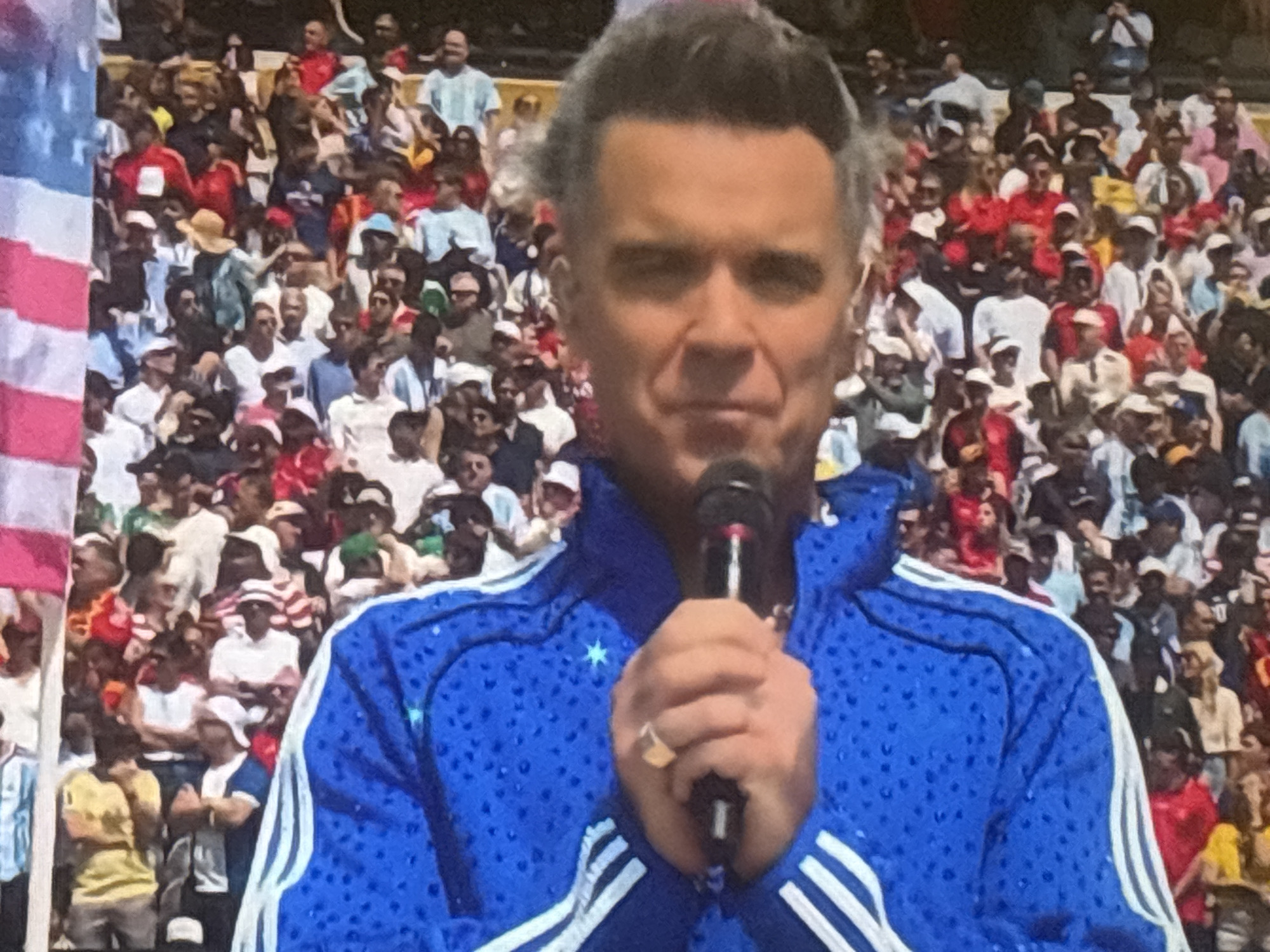 The image captures a live performance by Robbie Williams at the Lazur Stadium in Burgas, Bulgaria. Williams, a Caucasian male with graying hair, is dressed in a vibrant blue tracksuit with white stripes. He is holding a microphone and appears to be mid-song, with a ring visible on his left hand. The stadium is filled with a diverse crowd of spectators, many of whom are wearing casual clothing, and some are sporting hats and sunglasses. The atmosphere suggests a large-scale outdoor event, likely a concert or sporting occasion. The background is filled with rows of people seated in stadium stands, indicating a significant audience. The image is a still frame, possibly from a broadcast or recording of the event.