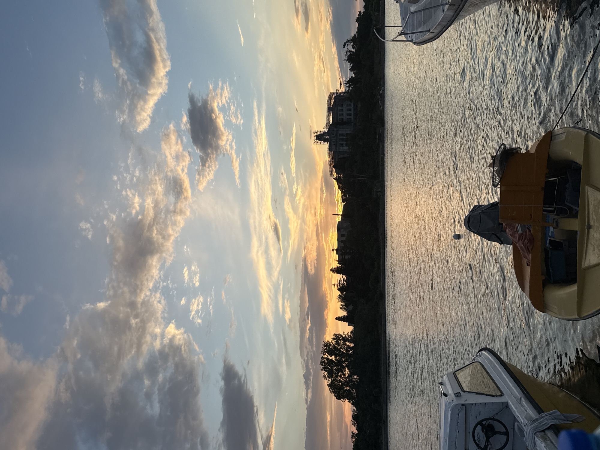 The image depicts a tranquil waterfront scene at sunset in Sozopol, Bulgaria. The sky is a canvas of soft blues, oranges, and yellows, with scattered clouds catching the fading sunlight. The water reflects the vibrant colors of the sky, creating a shimmering, ethereal effect. In the foreground, two small boats are moored, their forms silhouetted against the water. One boat is a white and yellow vessel with a steering wheel visible, while the other is a smaller yellow boat with an outboard motor.
Across the water, a tree-lined shore is visible, with a prominent building under construction perched atop a hill. The building, with scaffolding surrounding its upper levels, is silhouetted against the bright sunset. The overall atmosphere is one of peace and serenity, capturing the beauty of a coastal sunset in Sozopol.
