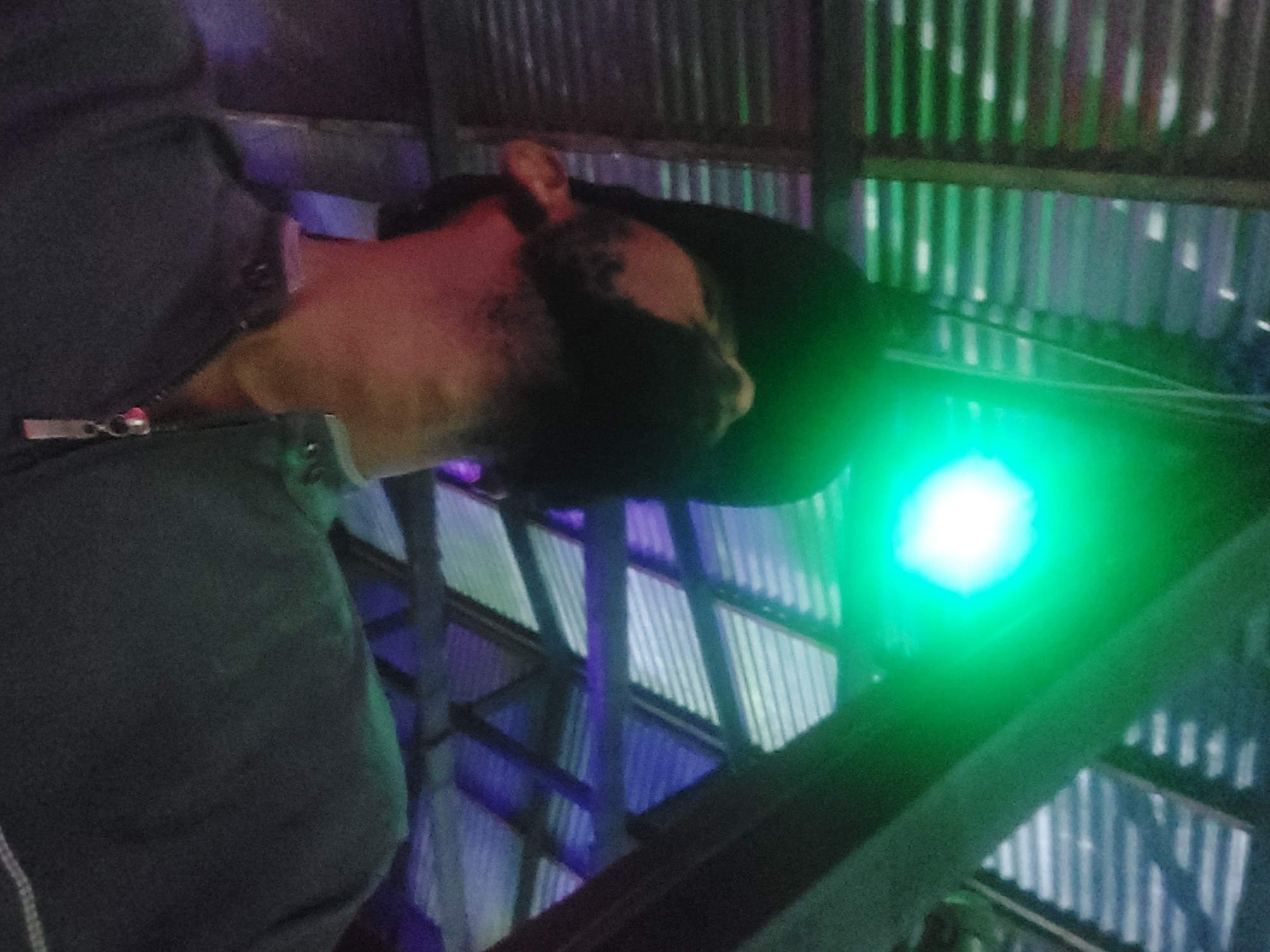 A man wearing a dark jacket and a black hat is seen from a low angle, illuminated by a green light. The background consists of corrugated metal and wooden beams, potentially a structure or building interior. The location is Dar es-Salaam, Tanzania.