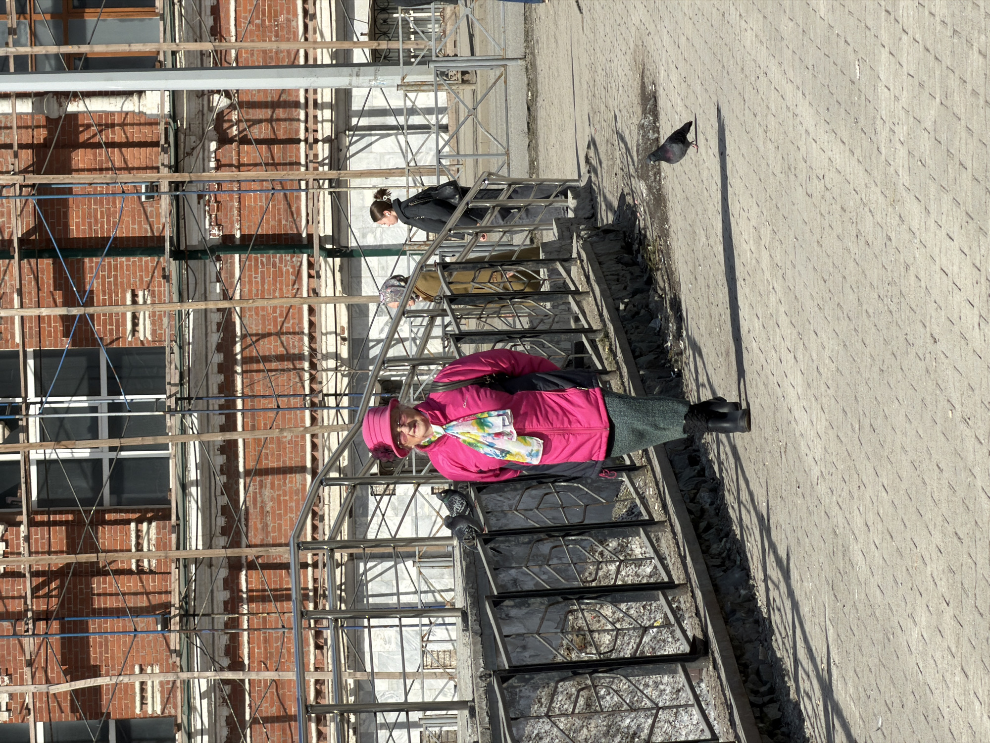 A sunny day in Perm, Russia, captures several individuals walking on a pedestrian bridge with scaffolding in the background. A woman wearing a bright pink jacket, pink hat, and patterned scarf stands prominently on the bridge. Other people are also present on the bridge. A pigeon stands on the concrete sidewalk. A brick building with visible windows is behind the scaffolding.