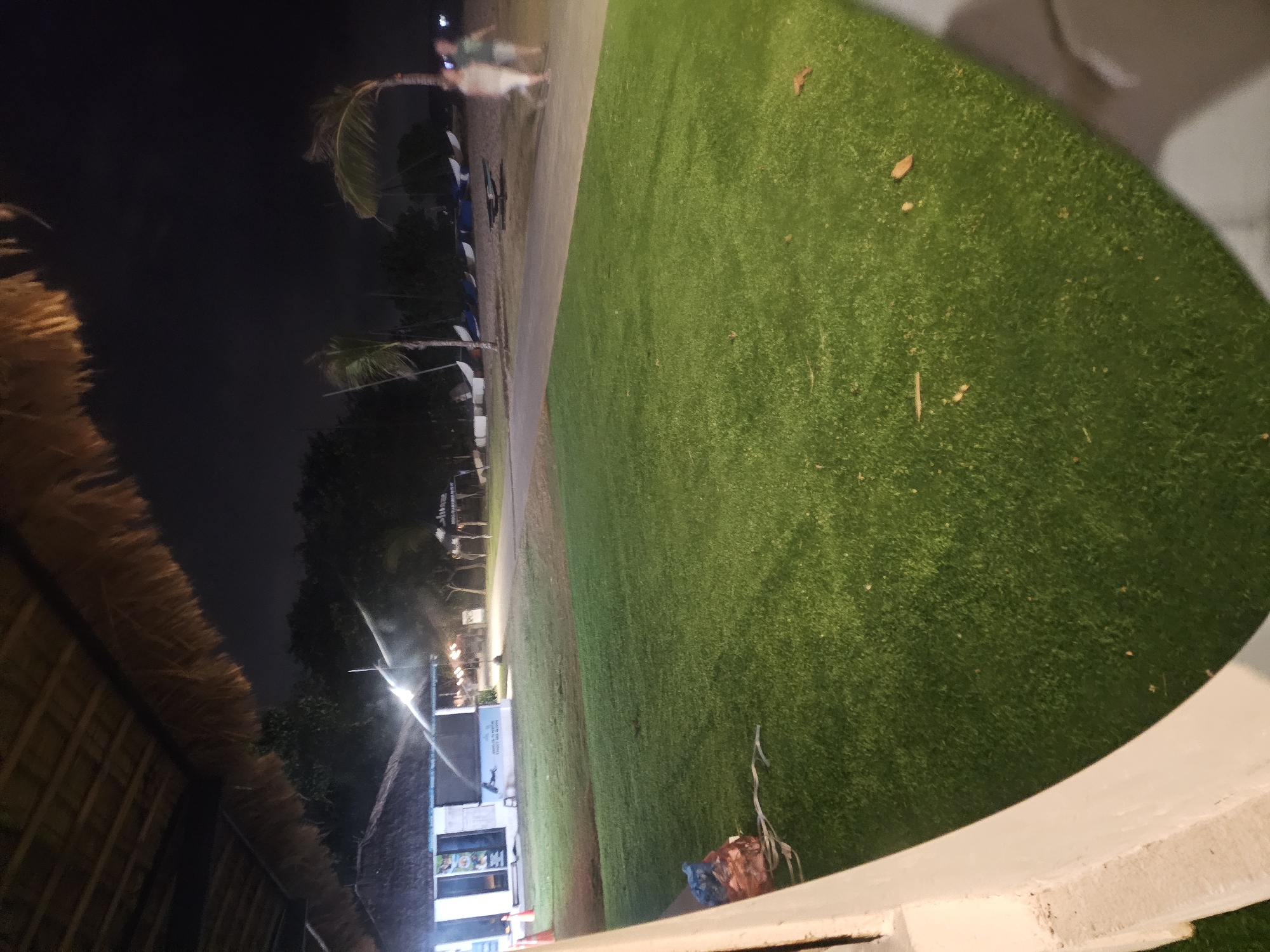 This is an outdoor night scene at a beachside location in Denpasar, Indonesia. In the foreground, there is a large expanse of manicured green grass. To the left, a white concrete wall or railing is visible. In the background, beyond the grass, there is a sandy beach area with several small sailboats lined up. Two palm trees are present, one on the beach and another more distant one. Further back, there are some trees and a building that appears to be a cafe or restaurant, illuminated with lights. Two individuals, appearing as blurs due to movement and low light, are walking on the sandy area near the sailboats. One person is wearing a light-colored top and trousers, and the other is wearing a dark top and shorts. The sky is dark, indicating it is nighttime.