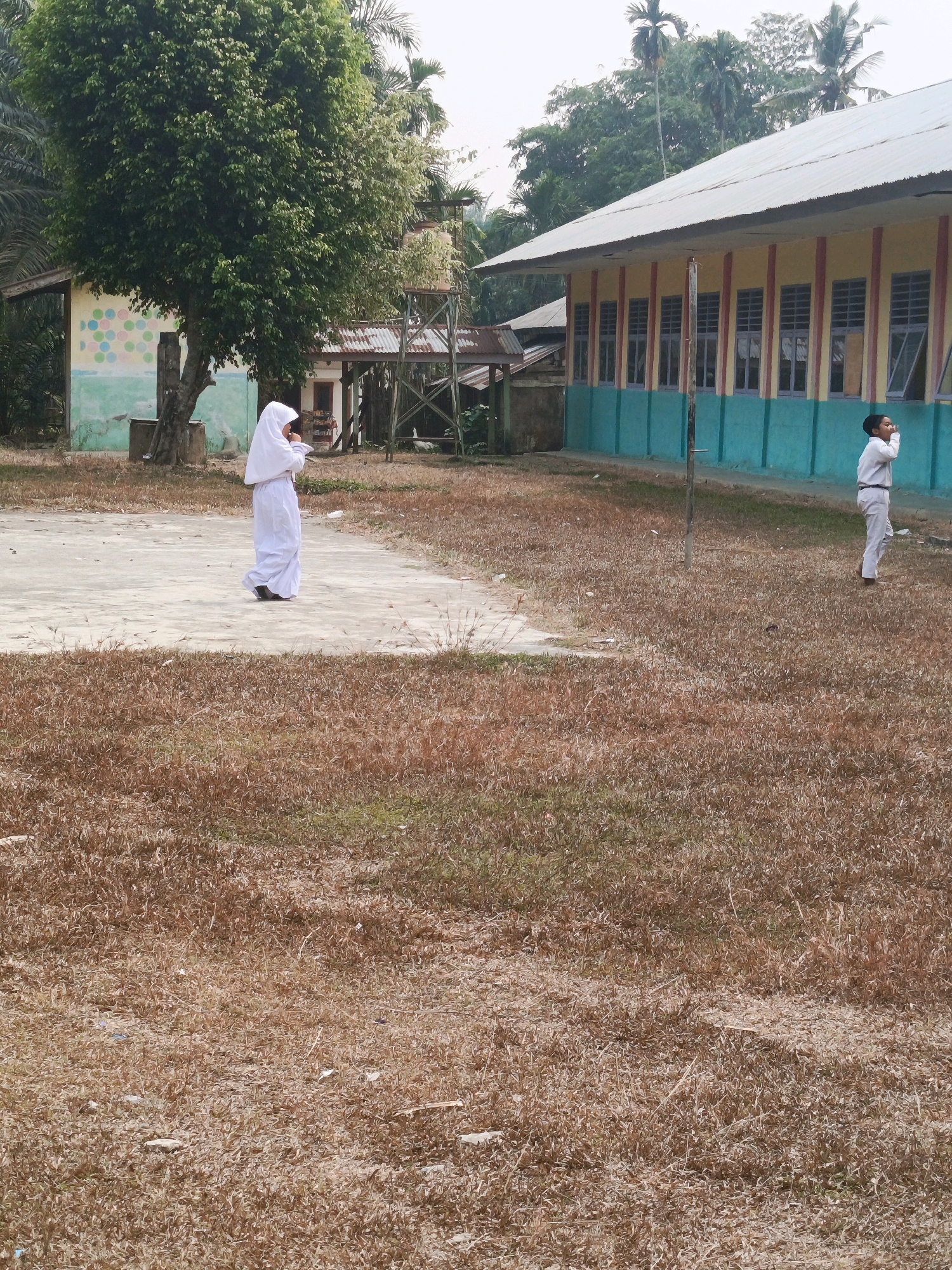 Two children are standing in a dry, grassy area in front of a yellow and teal building with a silver roof. The child on the left, wearing a white hijab and dress, is looking down at something held in their hands. The child on the right, wearing a white shirt and pants, is looking up and appears to be speaking or shouting. A large tree with green leaves is to the left of the children, and palm trees are visible in the background. The ground is covered in dry, brown grass. The building has multiple windows and a covered walkway. The image is set in Wonosari, Indonesia.