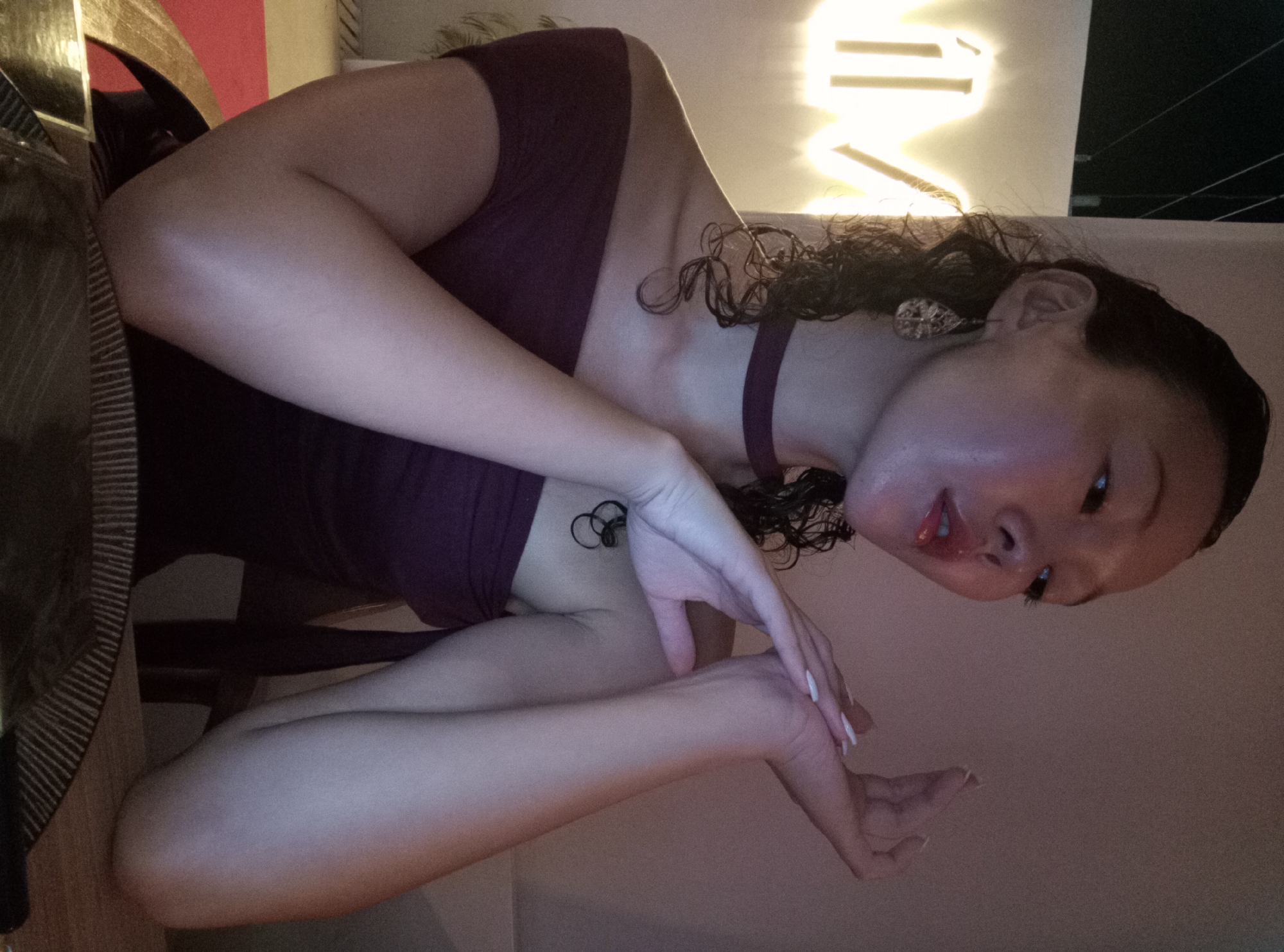 A woman with wet, curly dark hair and light skin sits in a low-lit interior. She is wearing a dark purple top with a matching strap around her neck. She has an earring in her left ear. Her left arm is extended and bent at the elbow, with her left hand near her right wrist. She looks down and to the right. The setting includes furniture like a table and chair. The location is presumed to be in North Kuta, Indonesia.