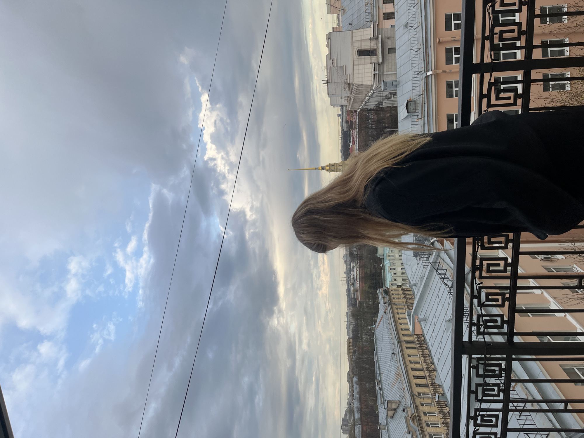 The image, taken from a high vantage point, shows a person looking out over a city from a balcony. The photo is rotated 90 degrees clockwise, making the horizon appear vertical.
In the foreground, the back of a person with long, straight blonde hair is visible, leaning forward over a dark, ornate metal balcony railing with geometric patterns. The person is wearing a dark-colored jacket or top.
Beyond the balcony, a sprawling cityscape unfolds under a partly cloudy sky. Numerous building rooftops, mostly with gray or metallic sloped roofs, are visible, indicating dense urban architecture. The buildings vary in color, including light brown, off-white, and grey. In the mid-distance, a distinctive golden spire, characteristic of landmarks like the Admiralty Building or the Peter and Paul Cathedral, rises above the rooftops. The presence of this spire strongly suggests the location is Saint Petersburg, Russia.
The sky is largely overcast with significant cloud cover, though patches of blue sky are visible, particularly towards the right side of the frame (as oriented in the original image). The light suggests daytime, possibly a late morning or afternoon with diffused sunlight. Thin wires or cables crisscross the upper part of the sky.
The scene conveys a moment of observation or contemplation of the urban landscape from above. There is no visible text in the image.