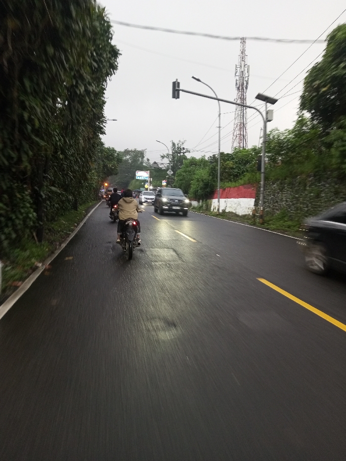 This image captures a street scene in the city of Dawathang_Dorjibi_ Kashingtsawa, Bhutan, under overcast daylight conditions. The paved asphalt road appears wet, reflecting ambient light.
The road features a solid white line on the far left edge and a solid yellow line marking the right boundary of the visible lane. Two distinct rectangular patches of darker asphalt indicate repairs on the road surface.
On the left side, a dense, tall wall of dark green foliage extends upwards, bordering the road. On the right side, a retaining wall painted with horizontal red and white stripes supports a vegetated embankment. Above the embankment, a tall, metallic lattice telecommunications tower is prominent against the gray sky.
Overhead infrastructure includes a street light fixture mounted on a pole on the right, equipped with a solar panel. Further down the road, a traffic light assembly hangs from an overhead gantry. Multiple utility lines traverse the upper portion of the frame.
Traffic is present in both directions. In the foreground, a person wearing a light-colored jacket is riding a dark motorcycle away from the camera in the left lane, with the red taillight illuminated. At least one additional motorcycle is visible behind the primary motorcycle. In the opposing lane, a dark-colored SUV with illuminated headlights is approaching. Other vehicles, including cars and motorcycles, are visible further in the distance. A dark vehicle appears blurred in motion on the far right edge of the foreground, moving from right to left.