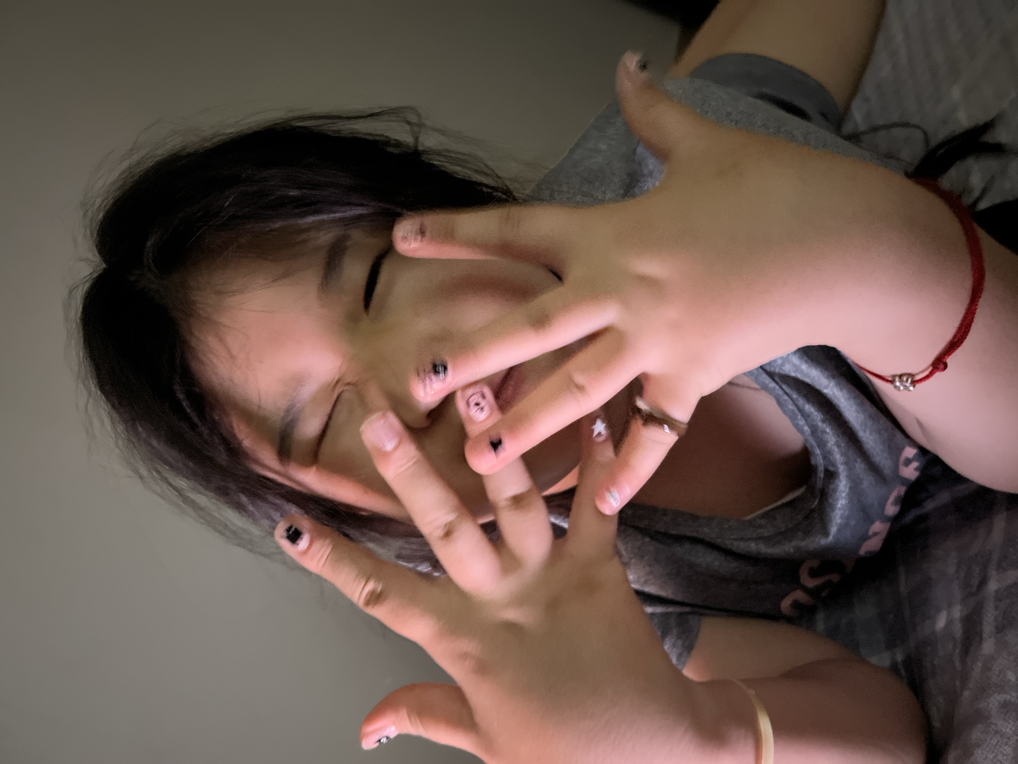 The image depicts a person with dark hair covering their face with both hands. The person is wearing a grey shirt, and a red bracelet is visible on one wrist. The fingernails are decorated with various designs, including black squares, a star, and a bear. The background appears to be a plain wall. The location and city are unknown.
