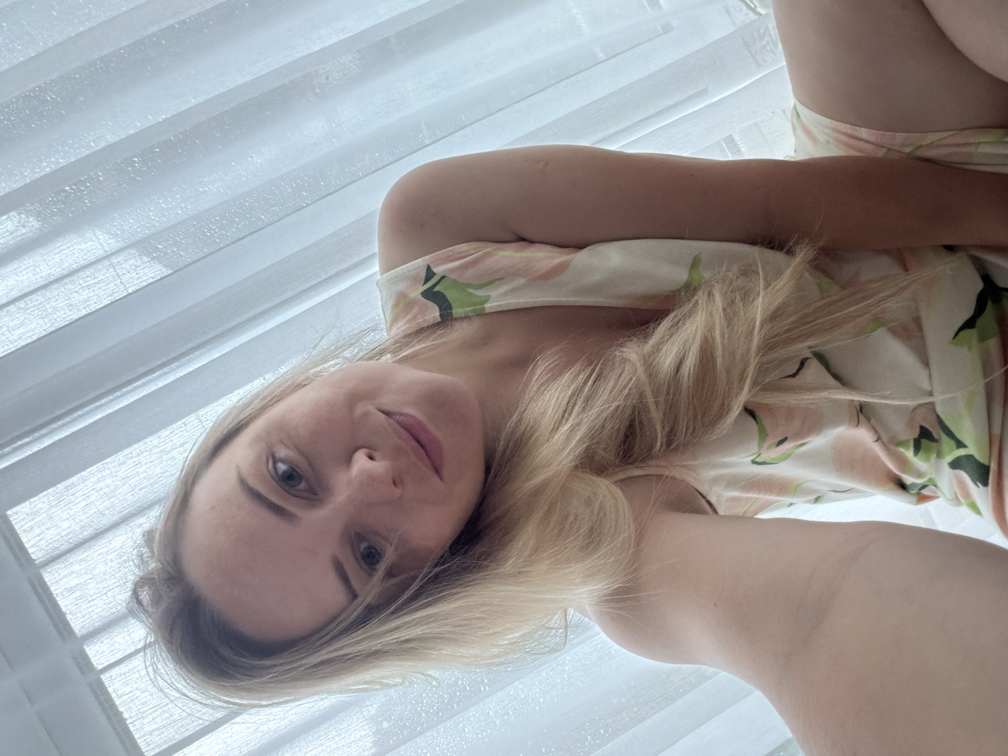A young woman with blonde hair and blue eyes smiles as she takes a selfie in front of a window with sheer white curtains. She is wearing a light-colored pajama set with a floral print. The scene appears to be indoors, likely a bedroom, with natural light filtering through the curtains. There are no identifiable landmarks or indications of Votkinsk, Russia.
