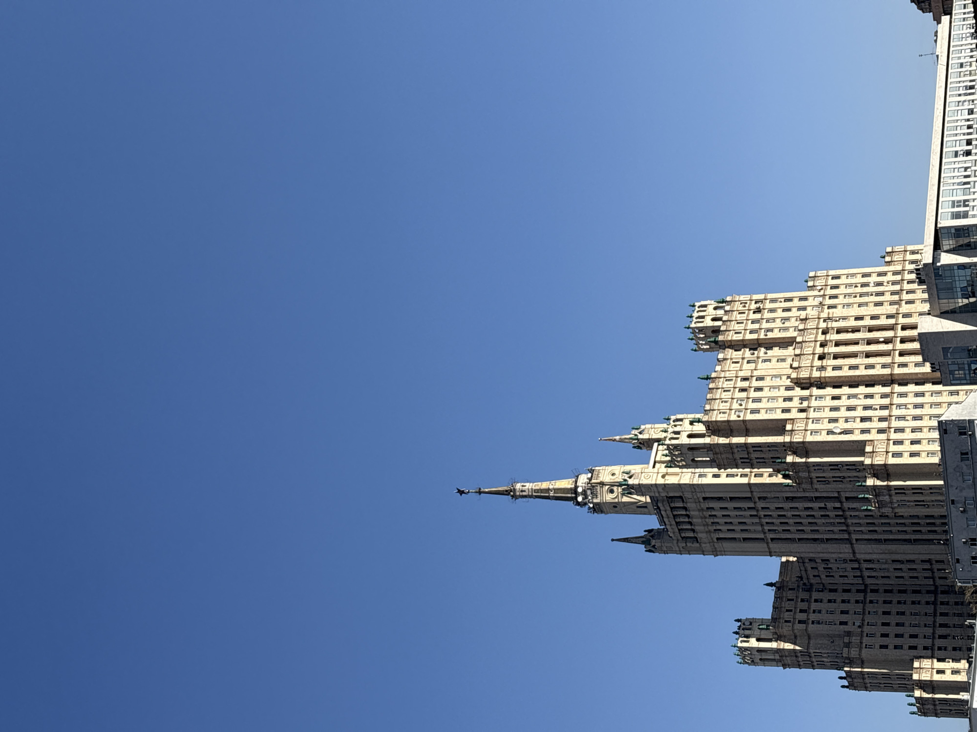 The image shows a Stalinist skyscraper in Moscow, Russia, viewed from below against a clear blue sky. The building's architectural style is characterized by its elaborate decorations, spires, and vertical emphasis. The structure appears to be well-maintained, with numerous windows visible on its facade. A modern building can be seen next to the Stalinist skyscraper.