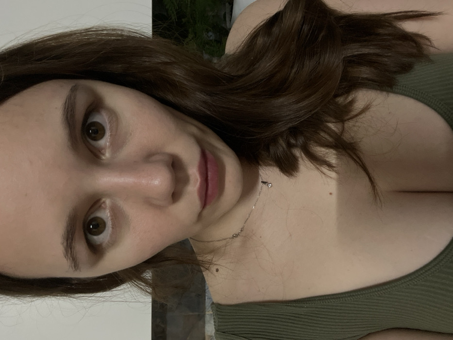 A light-skinned woman with brown hair and brown eyes gazes directly at the camera. She wears a green tank top and a delicate necklace. The lighting is soft, accentuating her features. The composition is a close-up, slightly angled. Background elements are blurry and indistinct, suggesting an indoor setting. The overall impression is intimate and personal.