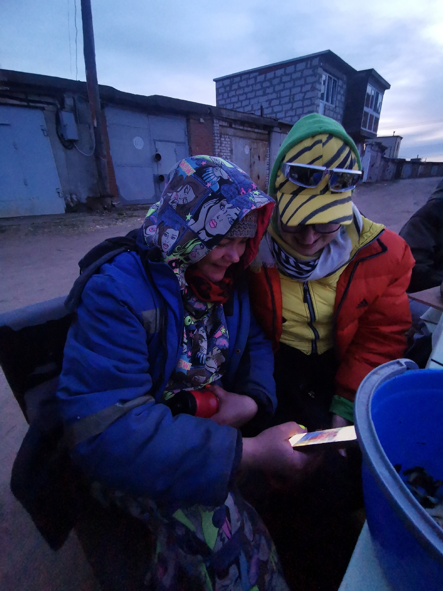 Two individuals are captured in an outdoor setting, engrossed in a smartphone during what appears to be dusk or dawn. The person on the left wears a blue jacket and a striking hooded sweatshirt featuring pop art-style female faces. A red scarf peeks from under the hood, and they hold a small red object while operating a bright smartphone with both hands. The phone's screen displays vibrant colors, including distinct blocks of red, blue, and yellow, illuminating their face.
The second person, to the right, is clad in a yellow puffer jacket beneath a red outer jacket, which features an Adidas logo on the sleeve. They wear a green hood with bold yellow and black stripes, and large mirrored sunglasses are perched on top of the hood. A black and white striped scarf is visible around their neck. Both individuals are leaning in, their attention fixed on the phone, suggesting shared interest or amusement.
The background consists of a row of grey industrial or storage garages with metal doors. One garage door displays a faded circular mark with the number "103". Above these garages, a small, two-story building constructed from cinder blocks with windows is visible. A utility pole stands vertically in the mid-ground. The sky is a muted blue-grey, indicative of low light conditions. In the bottom right corner, a blue bucket containing dark, unidentifiable items is partially visible. The scene portrays a moment of shared engagement in an utilitarian environment.