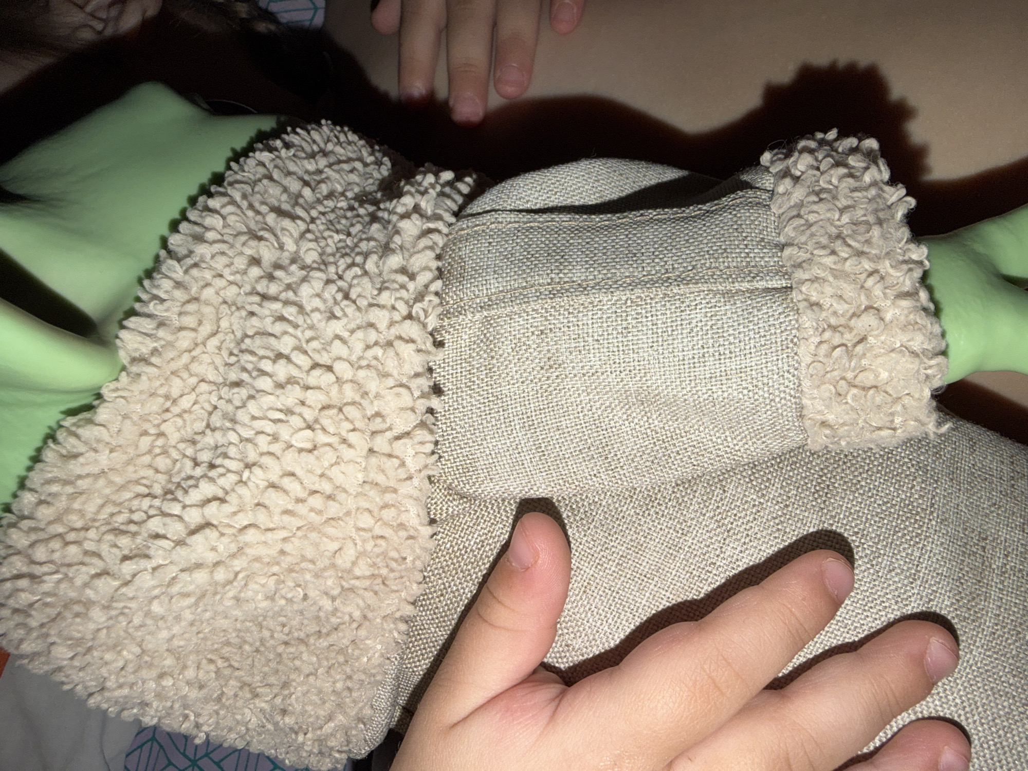 The image shows a close-up view of a plush toy resembling Yoda from the Star Wars franchise. The toy has large, light green ears with beige, curly-textured trim. Its body is covered in a beige, woven fabric. A child's hand is resting on the toy. Another child's hand is partially visible above the toy. The background is out of focus and indistinct. There is no indication that this image was taken in the city of Chaikovsky, Russia.