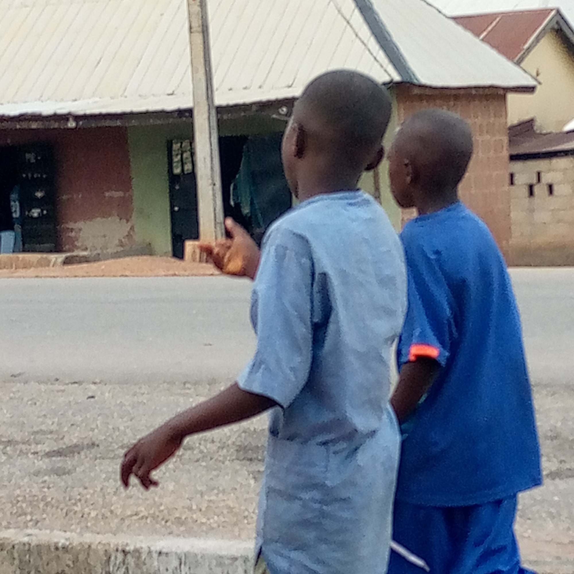 Two young boys are walking along a street in Mararaba, Nigeria. The boy on the left is wearing a light blue shirt and the boy on the right is wearing a dark blue shirt. In the background is a building with a white roof and green and brown walls. There is also a second building with a red roof visible in the background. The road is paved and there is a curb visible in the foreground.