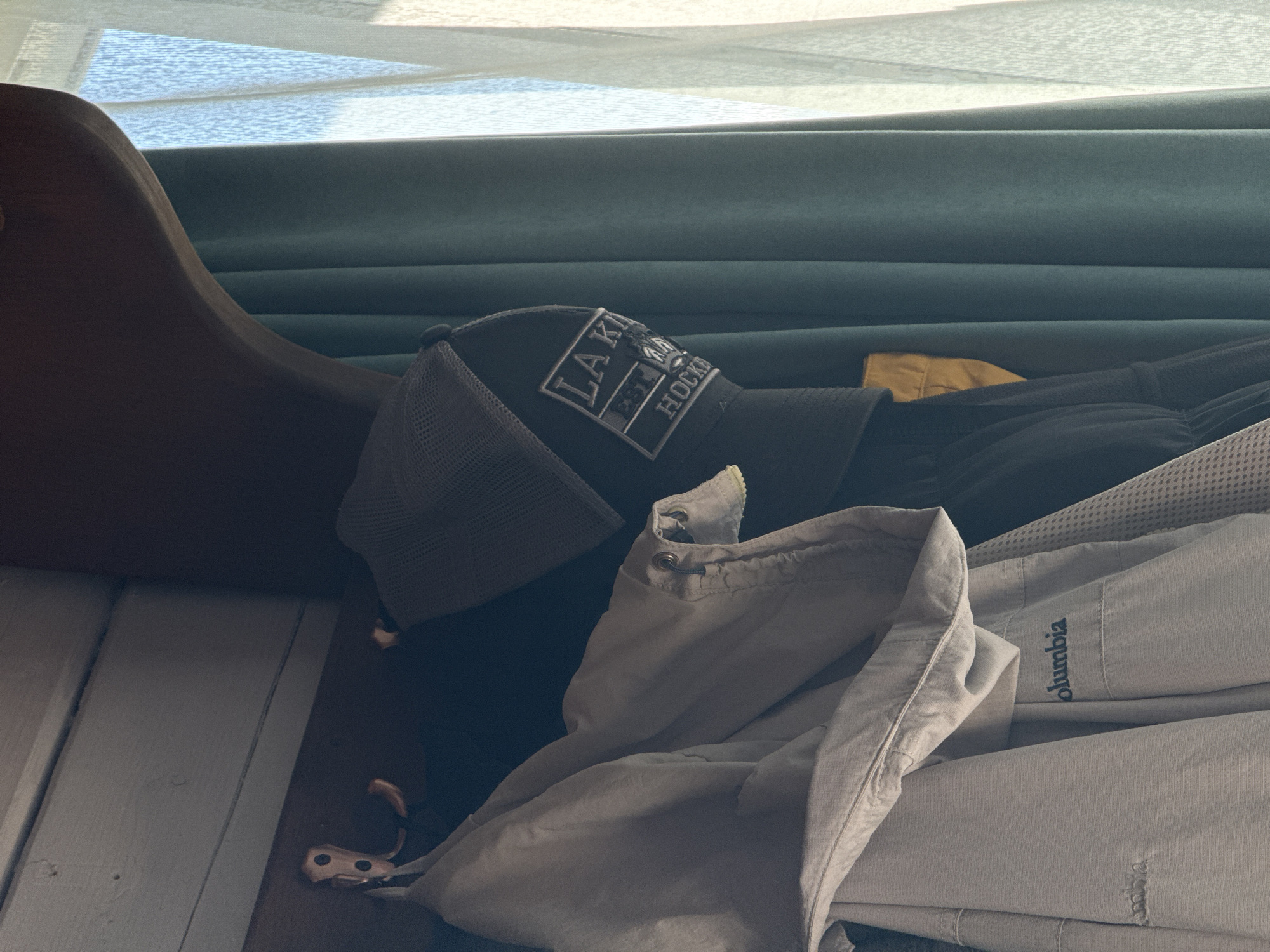 A close-up shot, taken from a slightly upward angle, shows several items of clothing hanging on hooks against a wooden panel. To the left, a light-colored jacket with the "Columbia" logo visible hangs on a hook. Next to it, a dark jacket or sweater is also hanging. On the right, a dark baseball cap with a patch reading "LA K EST. 1967 HOCK" is visible. The background features teal-colored curtains and a sheer white curtain, suggesting the items are hanging near a window, likely indoors. The lighting suggests it's daytime, with sunlight streaming through the window. The scene appears to be in a residential setting within the city of Artybash, Russia.