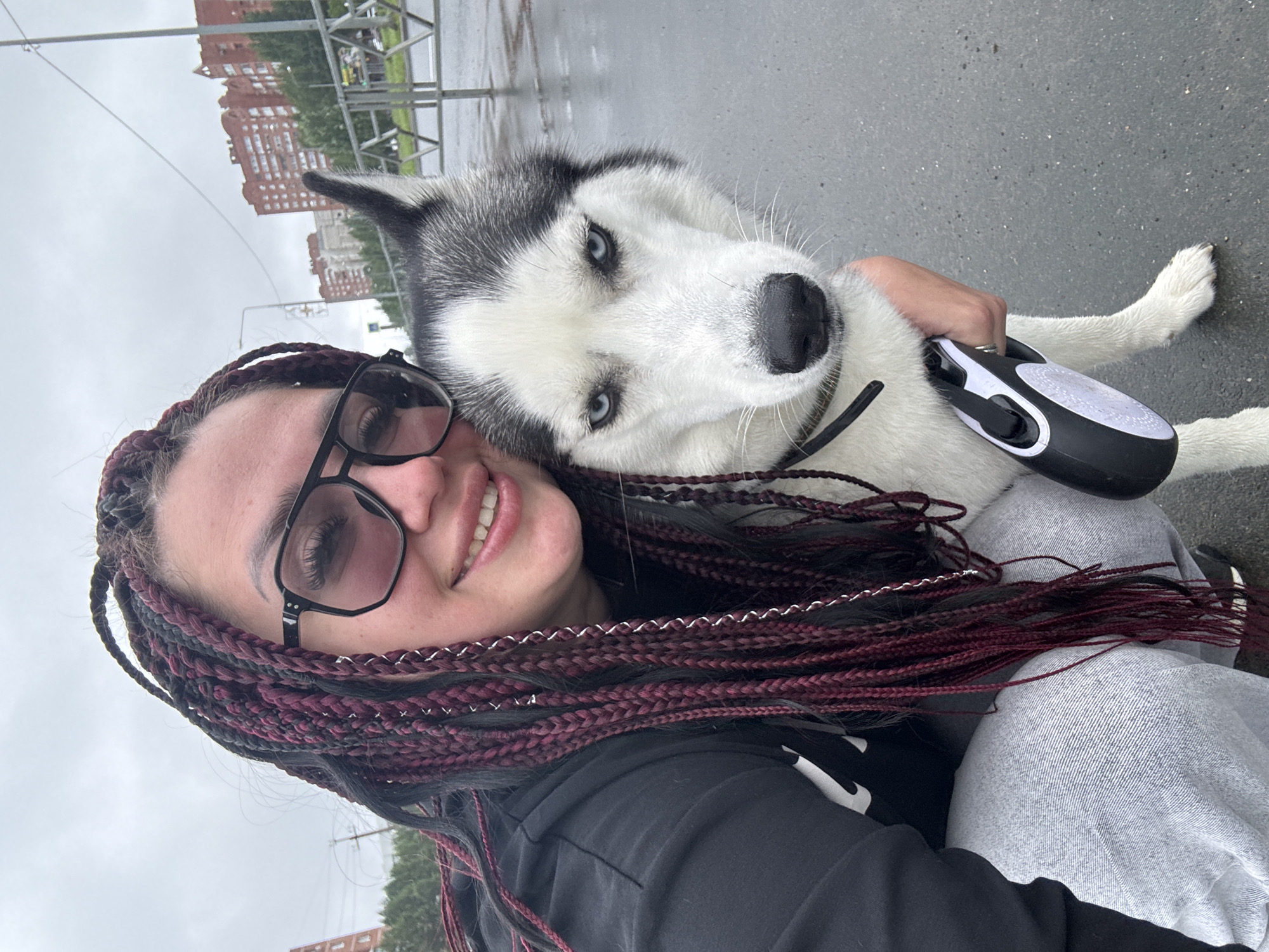 A close-up, eye-level, outdoor selfie shows a woman and a husky dog. The woman, positioned on the left, is wearing a black hooded sweatshirt and light grey sweatpants. She has dark hair styled in numerous long, burgundy and black braids, some with silver rings. She wears large, rectangular, black-rimmed sunglasses. Her right arm is around the husky, and her hand is holding a black and white retractable leash. The husky, on the right, has striking blue eyes and its head is tilted slightly towards the woman. The background depicts an overcast sky and a city street with red brick buildings and trees. The ground is wet, suggesting recent rain. The woman and dog appear to be in an urban park or on a sidewalk in Tolyatti, Russia.