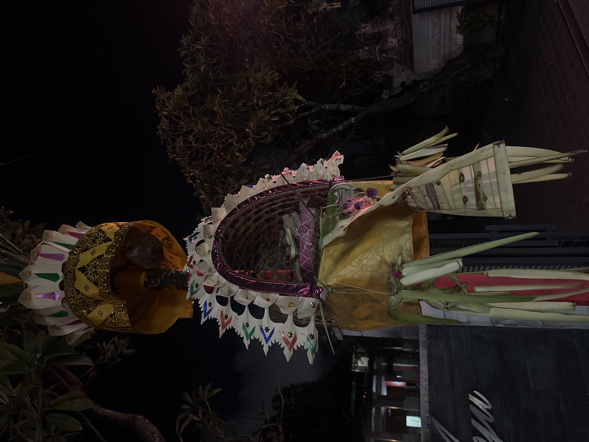 The image shows a religious offering, possibly a Penjor, a Balinese offering that is erected for ceremonies and festivals. It is an outdoor scene at night. The offering is tall and elaborate, decorated with woven palm leaves, fabric, and colorful paper. It hangs from a tree branch. In the background, there are trees, a building, and a paved walkway. The overall impression is one of cultural and spiritual significance within an urban setting in Denpasar, Indonesia.