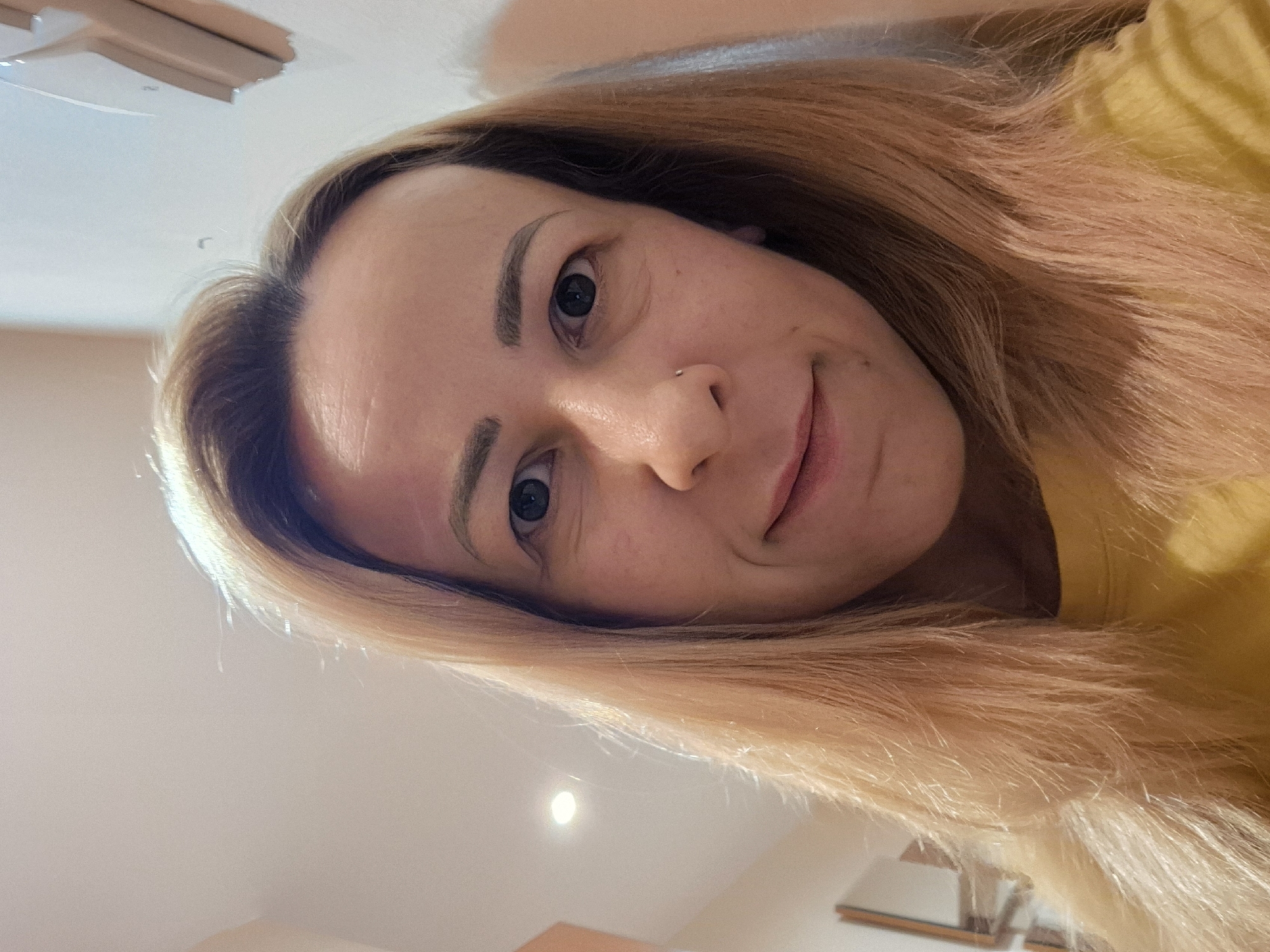 A medium shot captures a Caucasian woman with light brown hair, styled long and loose. She is facing forward, her head slightly tilted. Her gaze is directed towards the camera, and she has a subtle smile. A small silver stud is visible on her nose. The woman is wearing a yellow shirt. The background appears to be an indoor setting with a plain, light-colored wall and ceiling. A recessed light fixture is visible on the ceiling. The lighting is soft and even, suggesting an indoor environment. The overall impression is that of a selfie taken indoors.