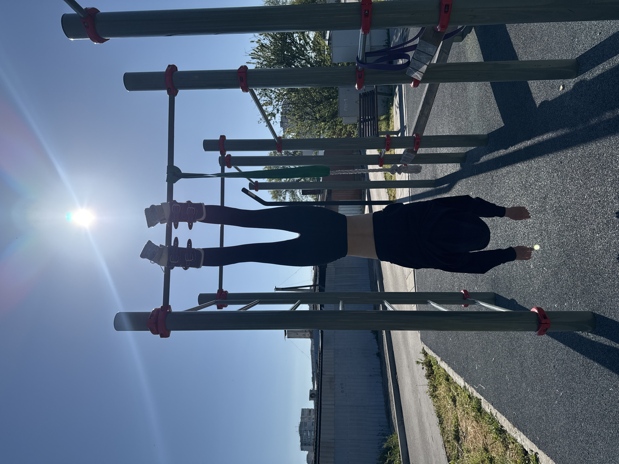 A person in black leggings and a black long-sleeved shirt is hanging upside down from a set of horizontal bars with their feet secured. They are outdoors on what appears to be a workout area with padded ground cover. The background features trees, buildings, and a blue sky with a visible sun glare. The location appears to be городское поселение Щелкино, Ukraine based on the cityscape.