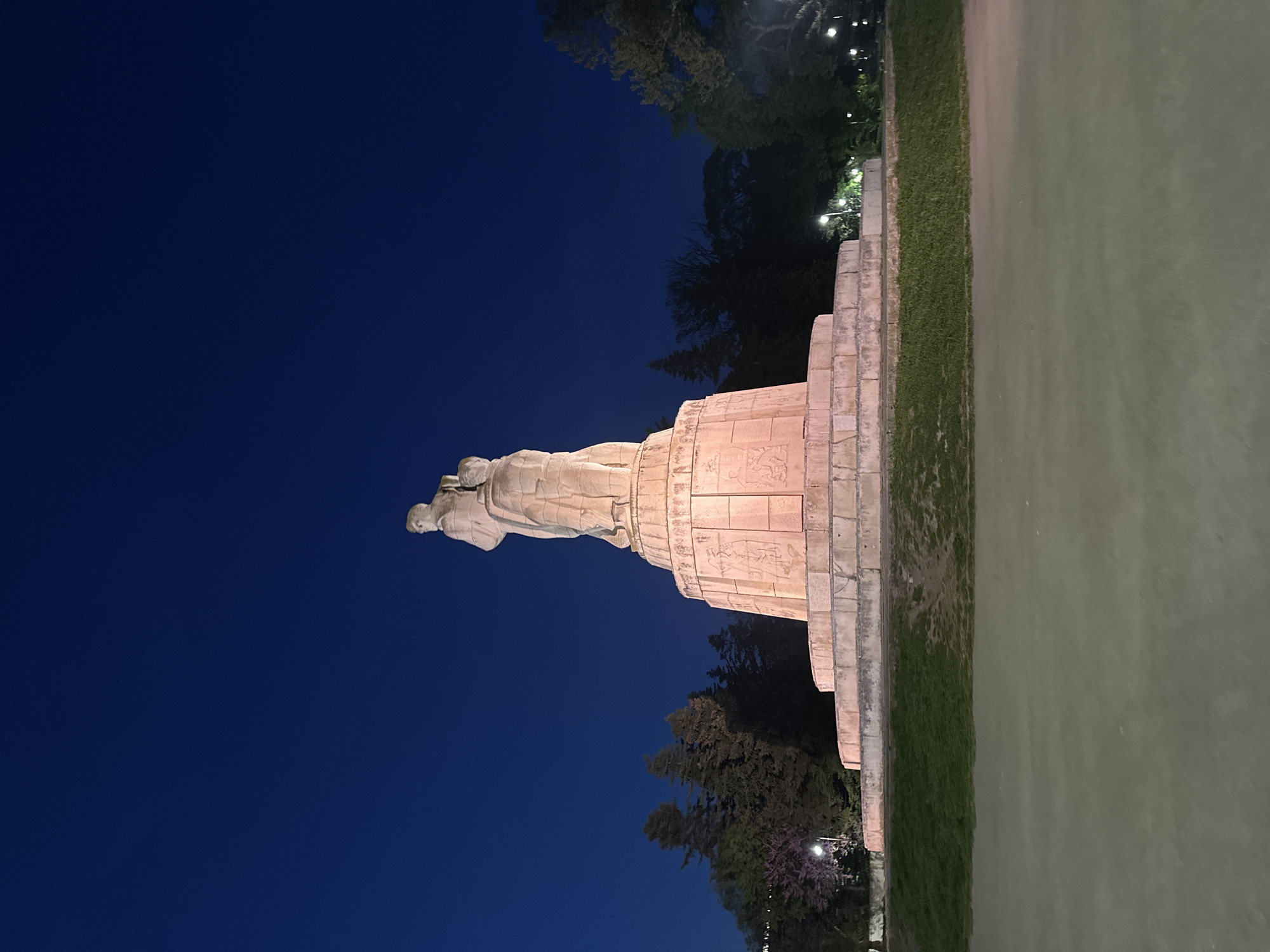 At night, a side view of the Monument of the Bulgarian-Soviet Friendship in Varna, Bulgaria is presented. The monument, constructed from light-colored stone, is adorned with reliefs and topped with a sculptural group of figures. The scene is illuminated by park lighting, revealing the structure’s details against the backdrop of the dark night sky and surrounding trees. A strip of greenery is adjacent to the monument, transitioning into an indeterminate light-colored surface.