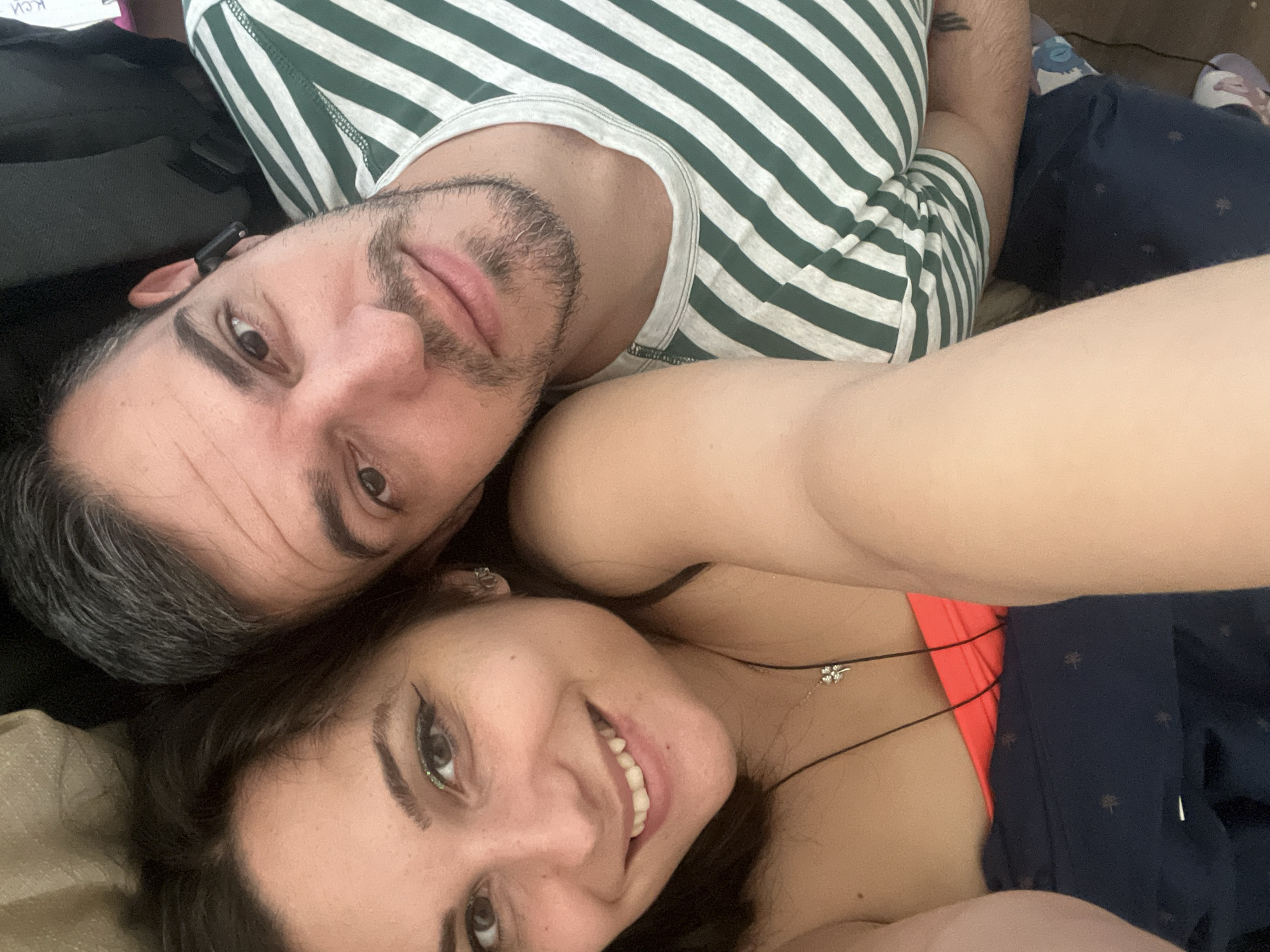A man and a woman are lying down, looking at the camera in a selfie. The woman is on the left, with her head tilted. She has dark hair, brown eyes, and is wearing a coral-colored top and a necklace. The man is on the right, with short, dark hair and a beard. He is wearing a green and white striped t-shirt and has an earbud in his right ear. They are in a casual, indoor setting. The background shows a blurred brown surface and a dark object, possibly a bag. There is no indication of the location in the city of Sofia, Bulgaria.