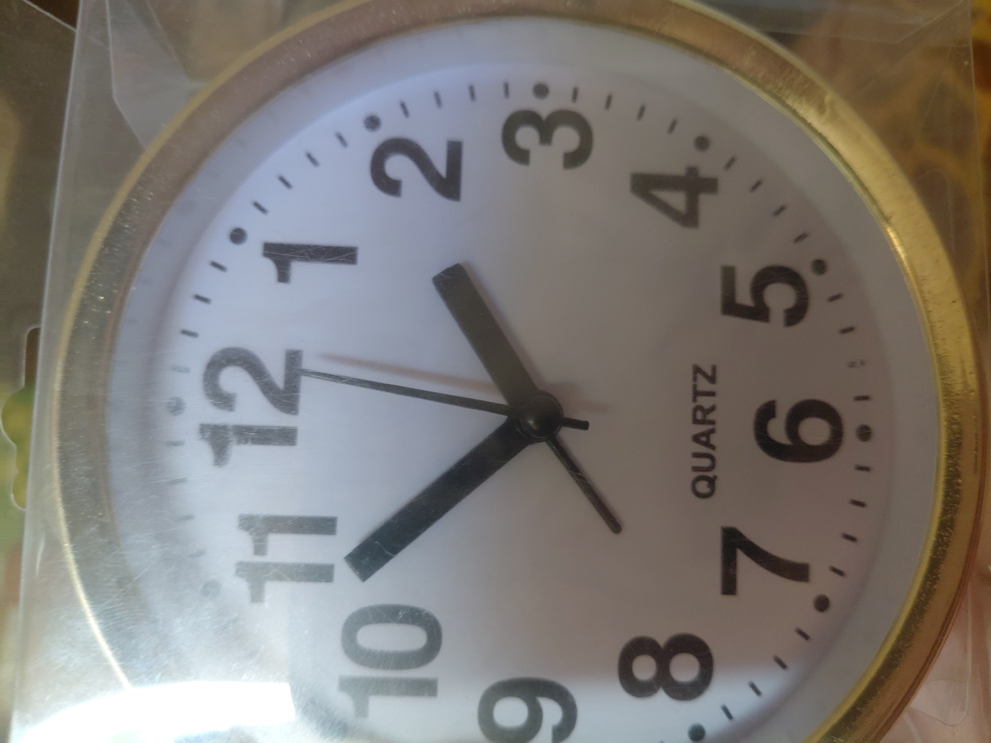 This is a close-up, slightly angled shot of a round analog clock with a gold-colored rim, possibly a wall clock or a clock that was part of another item. The clock face is white with black numerals and markers. The hour hand is pointing to approximately the 10 o'clock position, and the minute hand is pointing towards the 11 o'clock position, indicating the time is roughly 10:55. There is also a thin second hand visible, pointing towards the 5 o'clock position. The word "QUARTZ" is printed vertically in black text on the right side of the clock face. The clock is enclosed in clear plastic packaging, with some reflections and glare on the surface, suggesting it is new or being stored. The background is out of focus, appearing to be a cluttered indoor setting with warm, earthy tones. There are no people or animals visible. The scene is static, with no activity taking place beyond the movement of the clock's hands. The lighting is bright and somewhat diffused. The location context of Marynivka, Ukraine is noted, but there are no specific visual cues in the image to confirm this location.