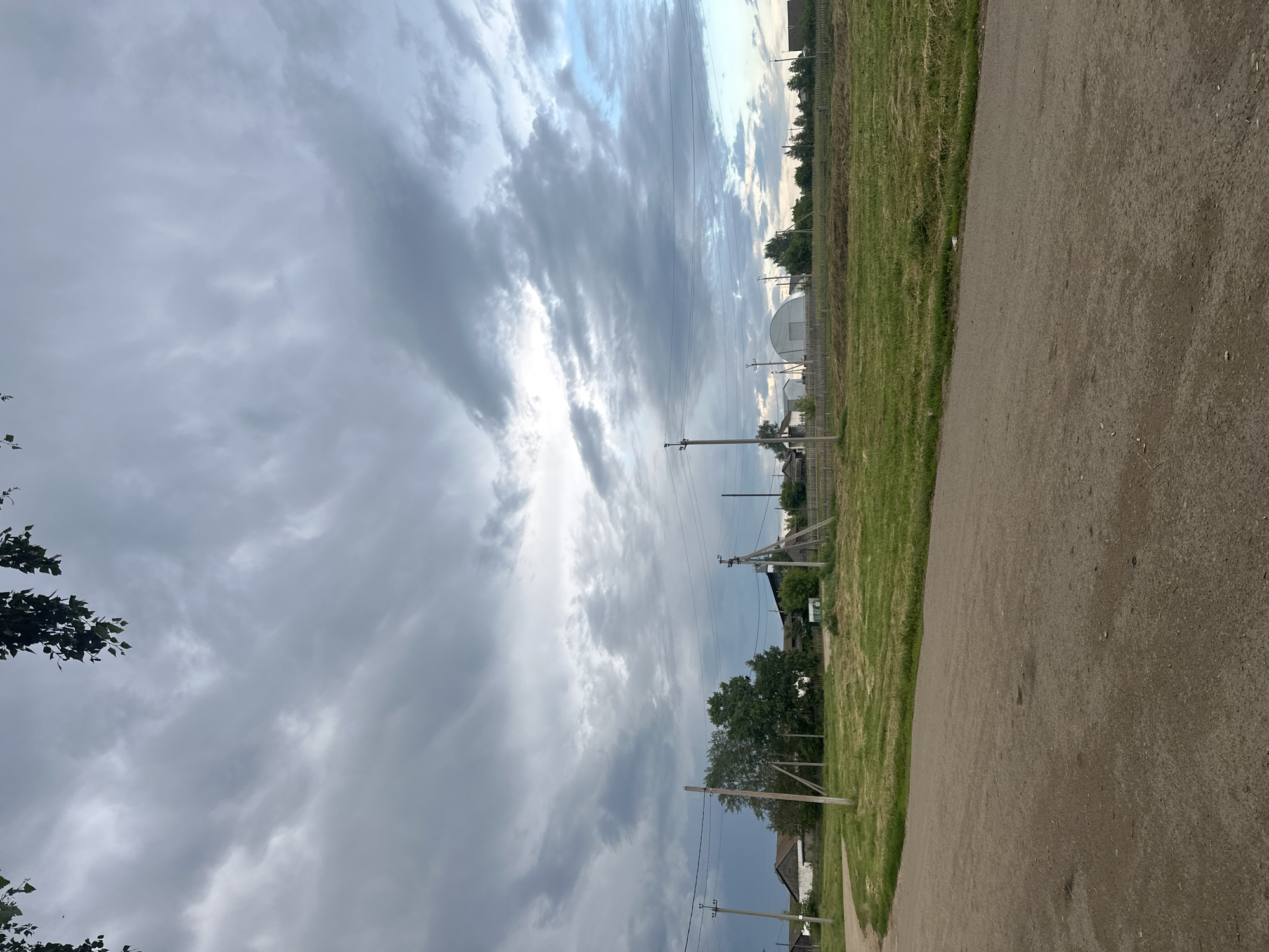 The image depicts a rural scene in Yaroslavka, Russia, on a cloudy day. A dirt road curves from the bottom left of the frame into the middle ground. To the left of the road, a grassy field stretches towards a small cluster of houses. Behind the houses, a line of trees is visible. In the middle ground, several utility poles with power lines traverse the scene. Further in the distance, a large, arched structure, possibly a greenhouse or agricultural building, stands out amongst more trees and houses. The sky is dominated by dramatic, grey, and white clouds, with patches of lighter blue sky visible towards the right. The overall impression is one of a quiet, overcast day in a village setting.