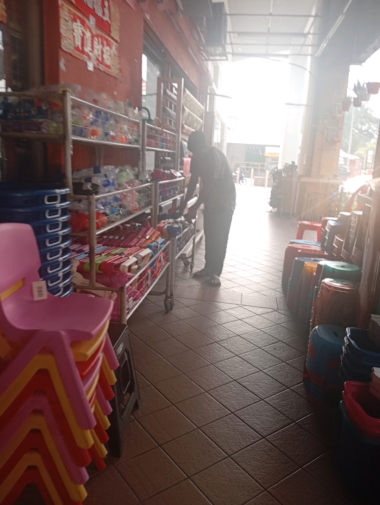 The image depicts a sun-drenched outdoor market stall located in the Selayang Municipal Council, Malaysia. The stall is filled with various items, including stacks of colorful plastic chairs in shades of pink, yellow, and red on the left side of the frame. Next to the chairs, metal shelving units are laden with an assortment of goods, many of which appear to be packaged drinks or food items in plastic bottles and wrappers. A person, seen from behind and in profile, is standing in the middle of the aisle, reaching towards the shelves, possibly arranging or selecting items. Further down the aisle, more shelves are visible, as well as rows of cylindrical stools in red, blue, green, and orange. The ground is paved with brown square tiles. The scene is illuminated by bright sunlight, creating strong highlights and shadows, suggesting it is daytime. In the background, a building with a covered walkway and some greenery can be observed. The overall impression is of a busy market environment.