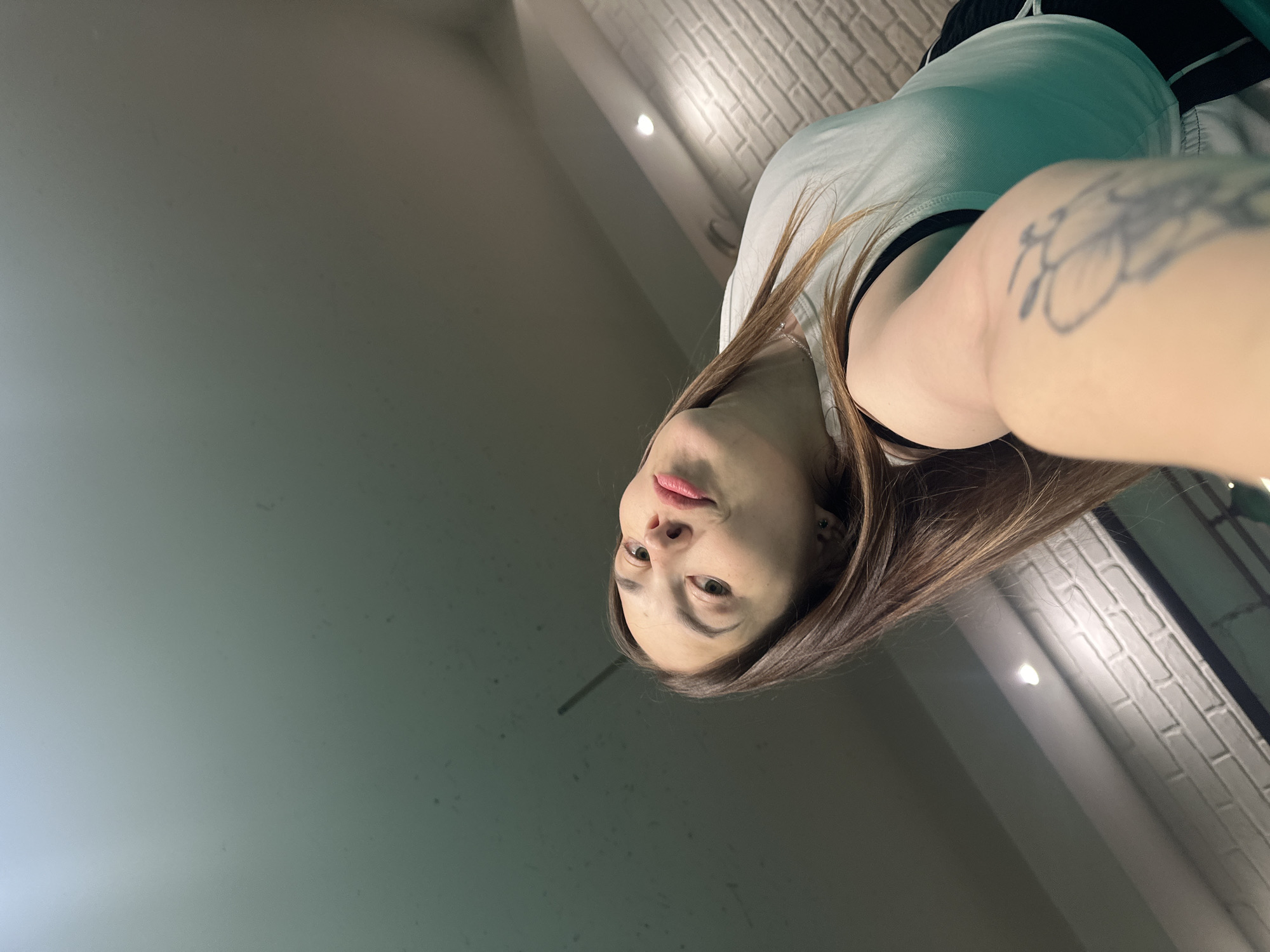 A young woman with long brown hair and bright pink lipstick is taking a selfie. She is wearing a white tank top and black shorts with white trim. A tattoo is visible on her left arm. The background appears to be an indoor setting with white brick walls and overhead lighting. There is no indication of the city of Perm, Russia, or any specific location details.