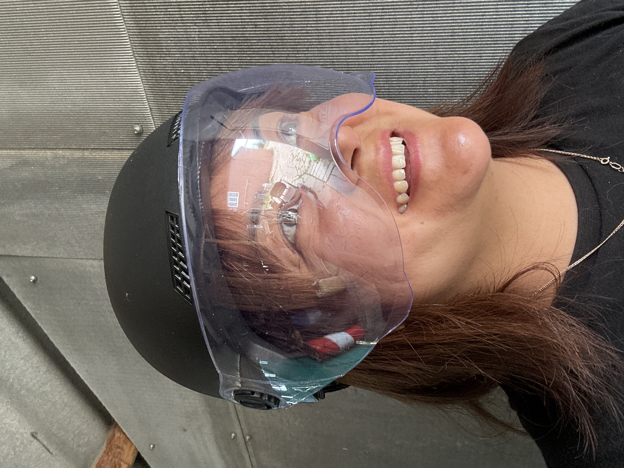 A medium close-up shot shows a woman wearing a black helmet with a clear visor, her face partially obscured by the visor. The visor reflects a building and a paved area. She has brown hair, is smiling, and appears to be outdoors, possibly in a garage or shed, given the corrugated metal wall in the background. She is wearing a black t-shirt and a delicate necklace.